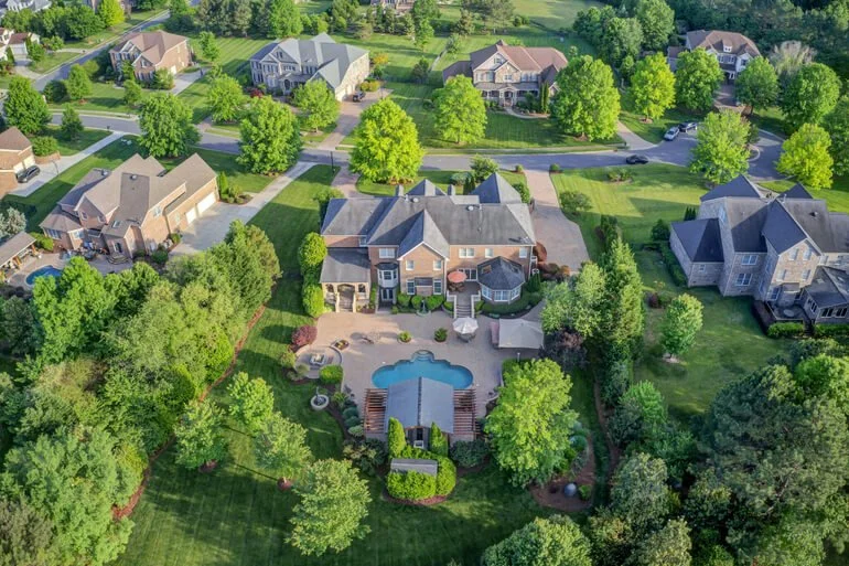 Aerial view of a suburban neighborhood with large houses, green lawns, and numerous trees. The central house features a backyard with a swimming pool, patio, and landscaped garden.