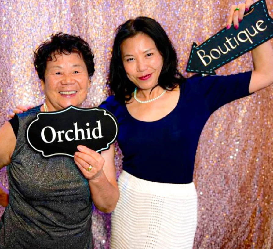 Two women smiling and holding signs, one says "Orchid" and the other says "Boutique". They stand in front of a sparkly pink backdrop.