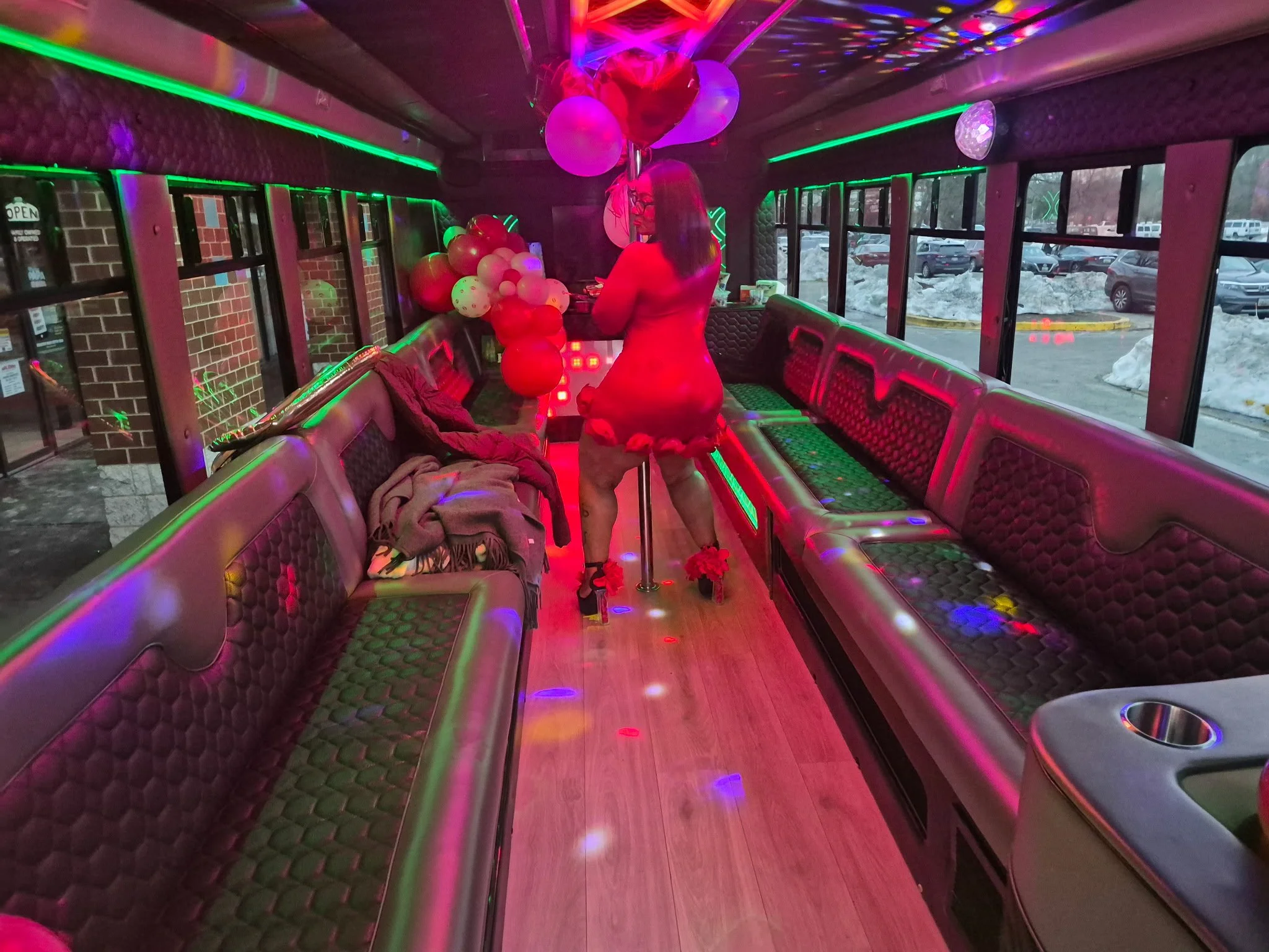 A woman wearing glasses and a red dress is dancing on a dance pole inside a party bus decorated with balloons. The bus has dim lighting with colorful LED lights, and through the windows, a snowy parking lot is visible.