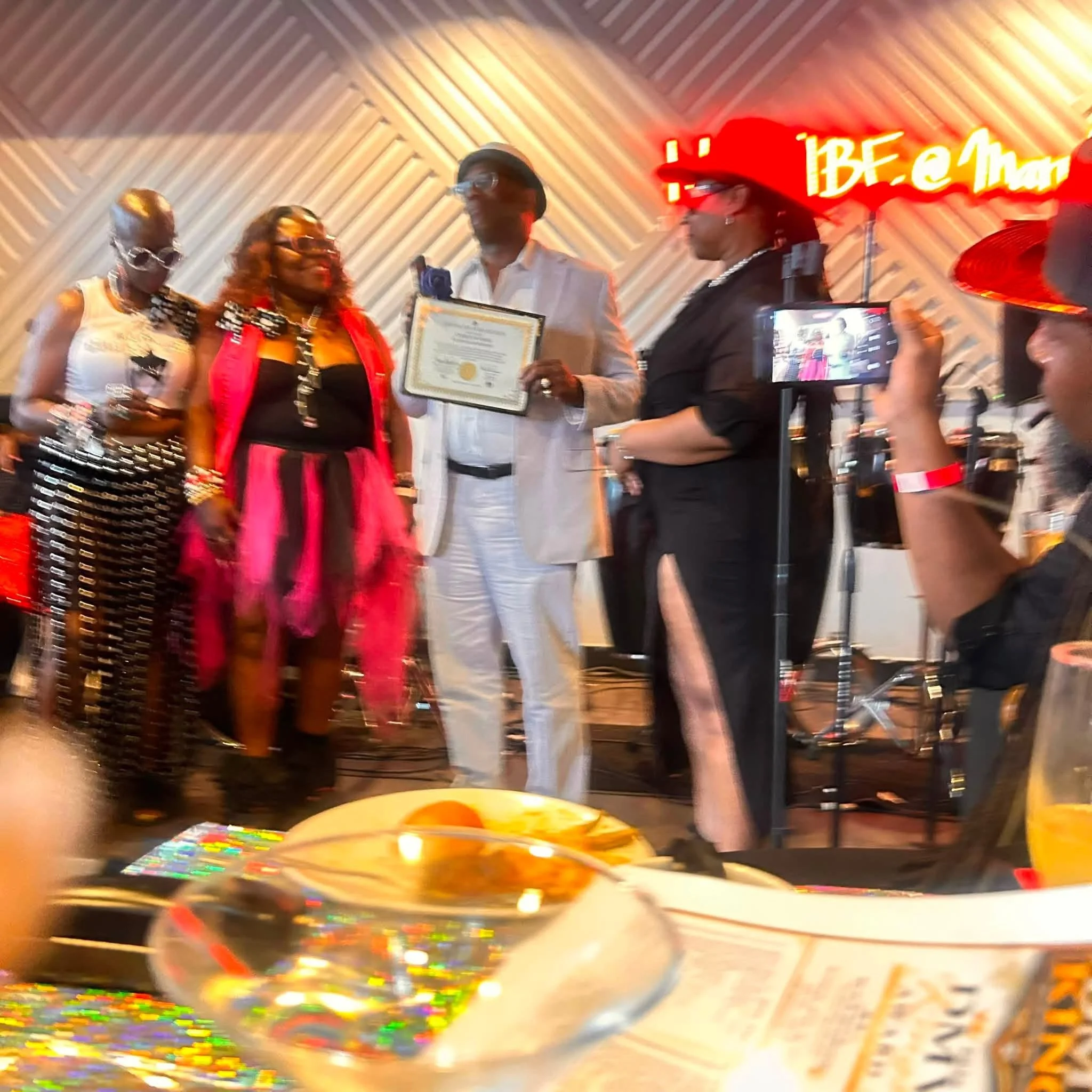People at a celebration gather around a table with colorful decorations, including balloons and a chocolate cake with rainbow sprinkles. In the background, a red neon sign says 'BE' and other words that are partially visible. One person is holding a 