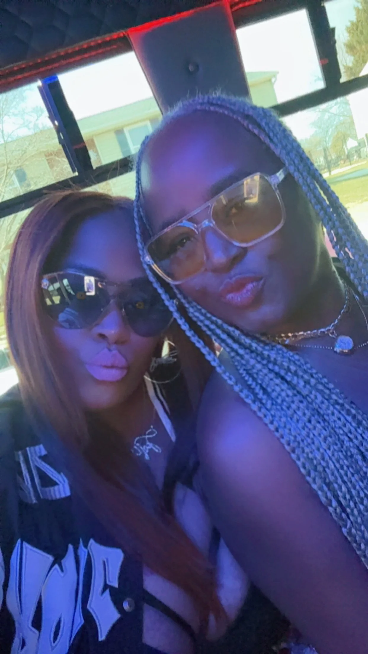 Two women take a selfie together outdoors, both wearing sunglasses and bold makeup, with one having long braided hair and the other with straight, shoulder-length hair.