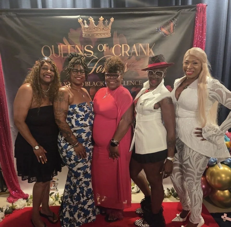 Five women standing in front of a backdrop that reads "Queens of Crank" with a crown graphic. They are dressed in colorful, fashionable outfits, smiling, at a celebration event.