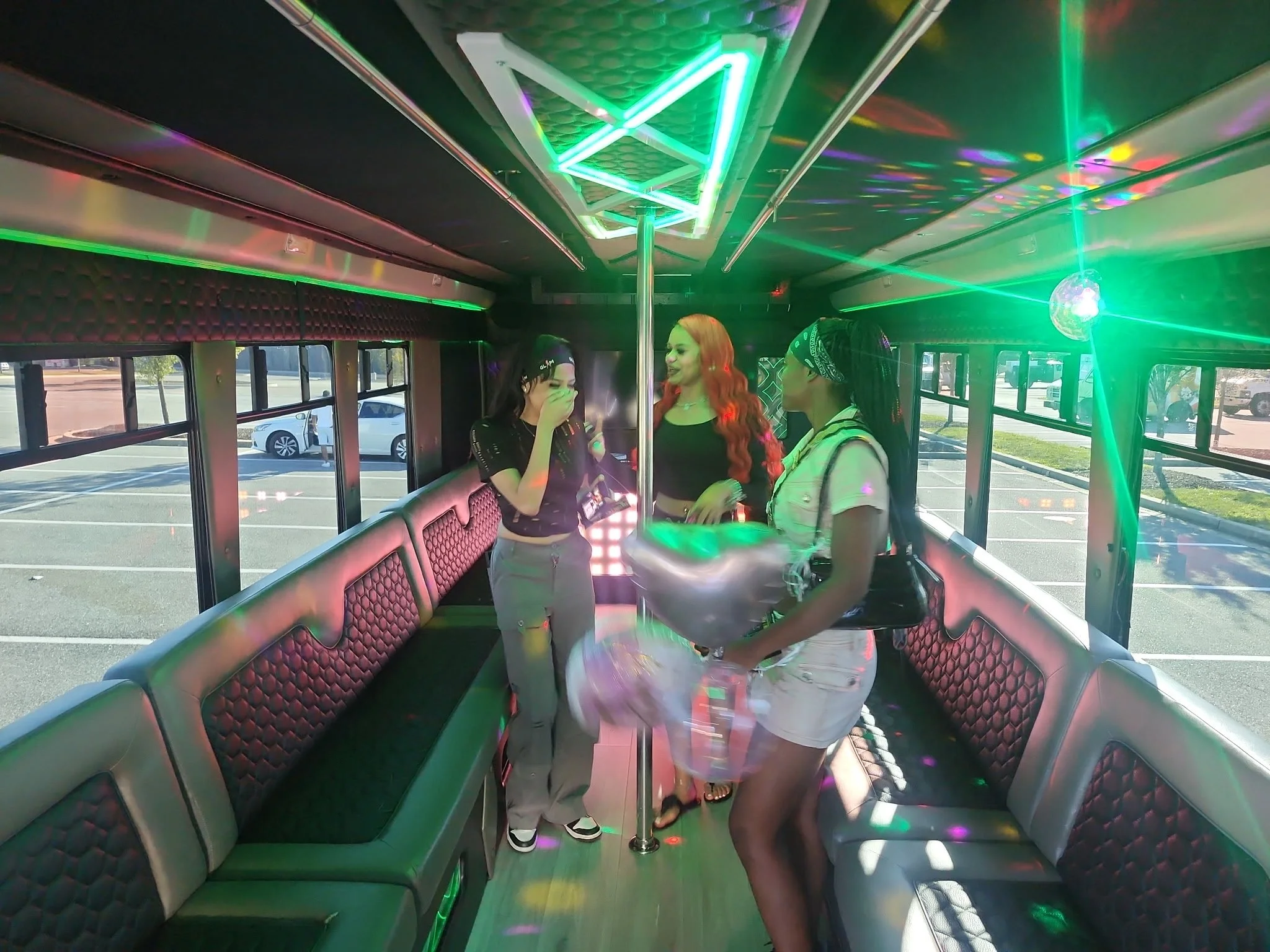 Inside a party bus with colorful neon lights, three women are standing near a pole, chatting and laughing, with a parking lot visible through the windows.