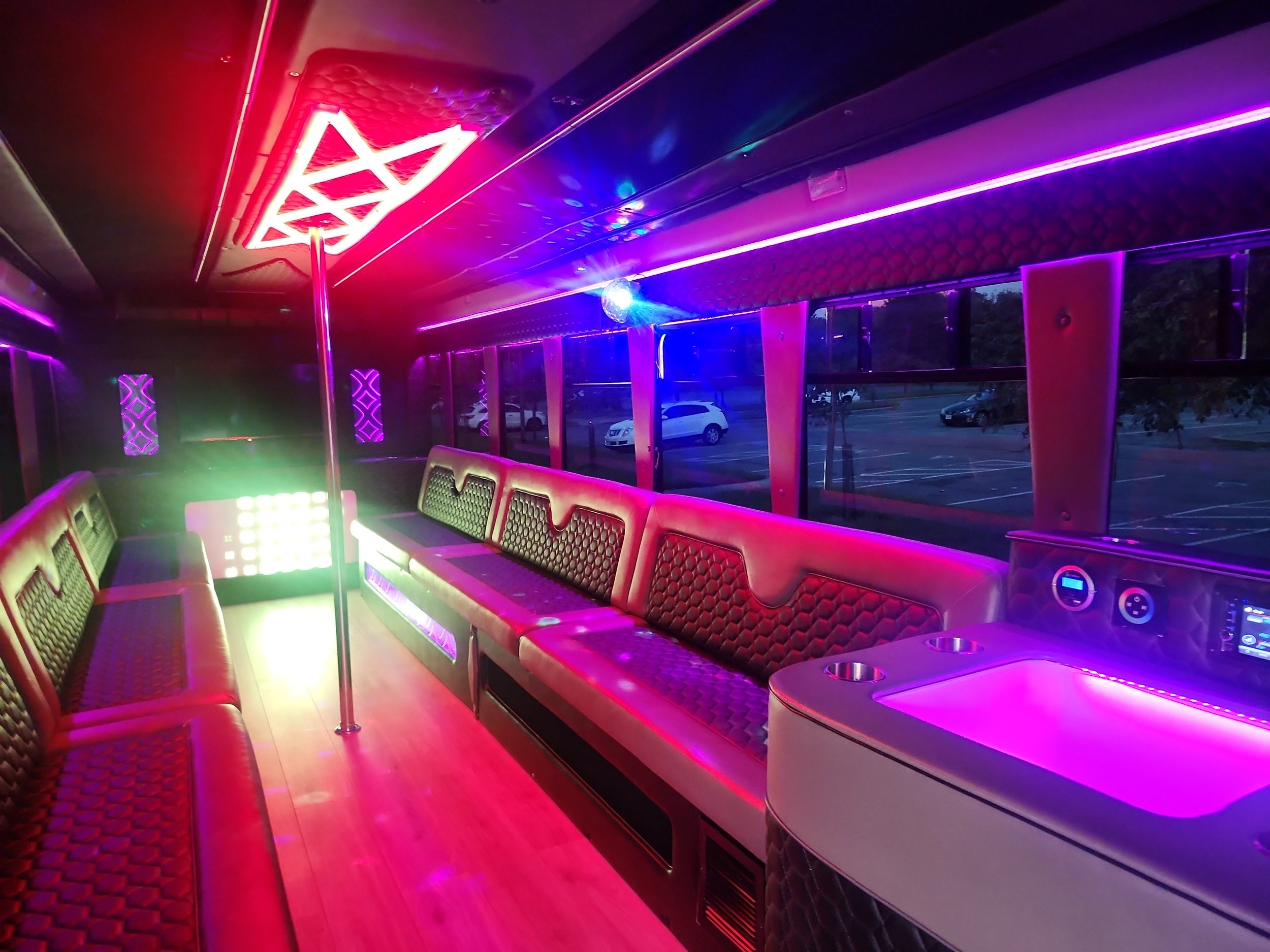Interior of a party bus with pink, purple, and blue neon lights, red cushioned seats, and a disco ball on the ceiling.