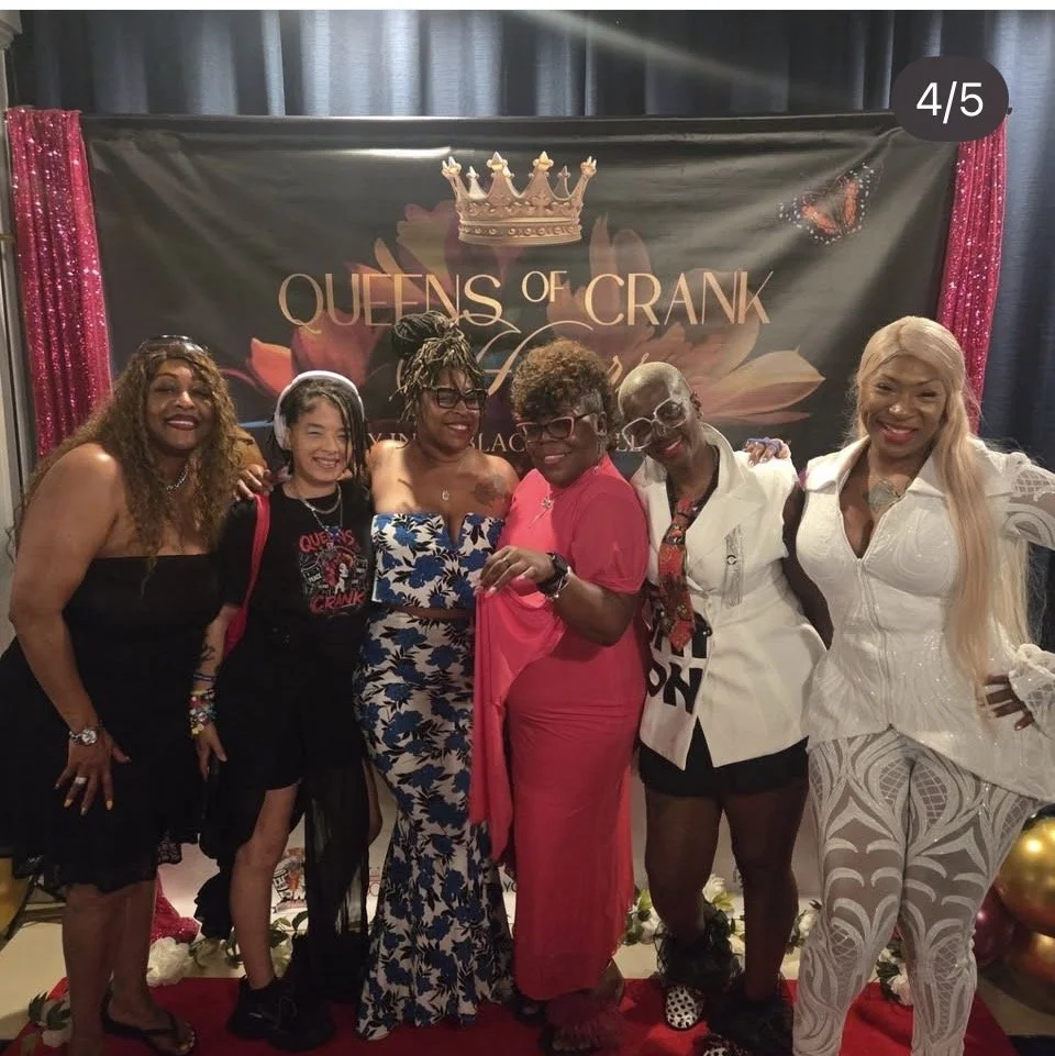 Group of six women standing together in front of a backdrop that reads 'Queens of Crank' with a crown graphic, pink and gold balloons at the bottom, all smiling and posing for the photo.
