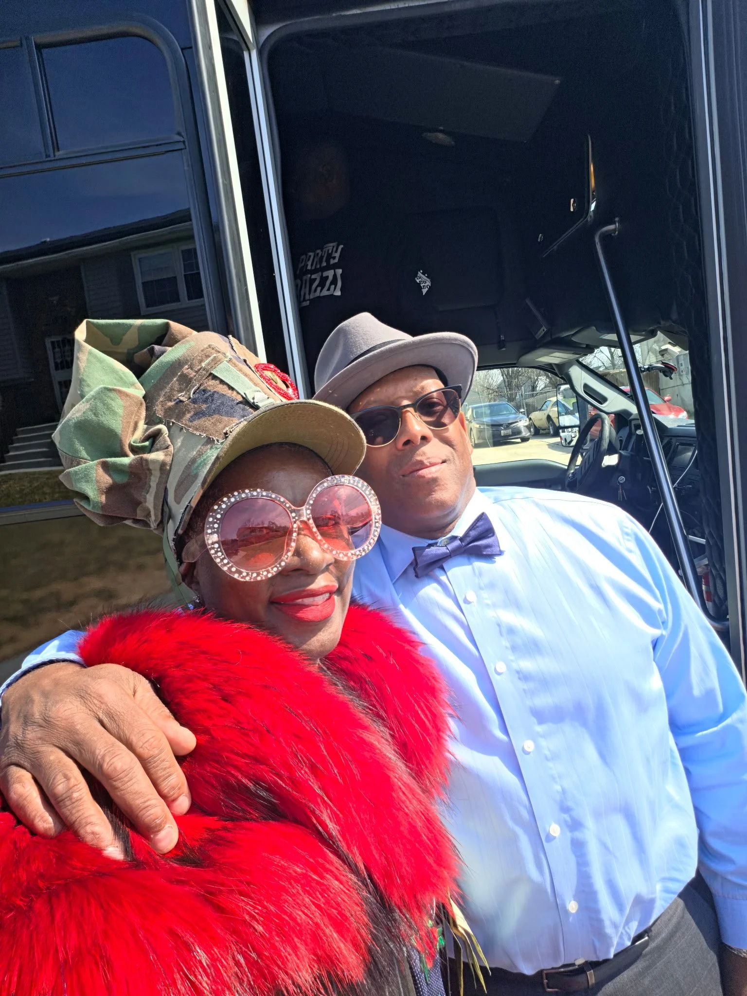 A man and a woman taking a selfie together outdoors. The woman is wearing a colorful hat, large heart-shaped sunglasses, and a red faux fur coat. The man is dressed in a light-colored dress shirt, bow tie, and sunglasses. They are smiling and appear 