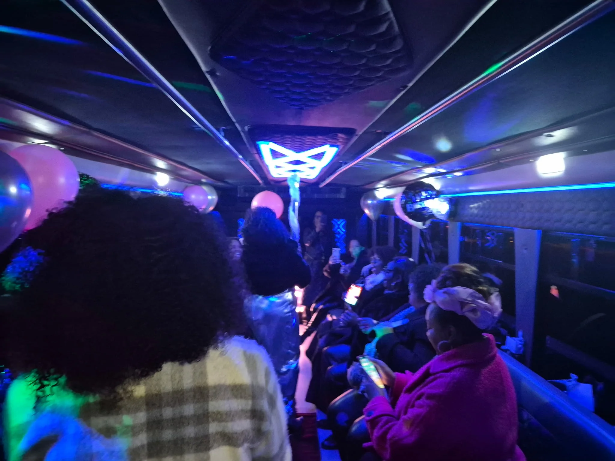 People sitting in a dimly lit room with neon lights, balloons, and a central neon logo or symbol on the ceiling, possibly at a party or nightclub.