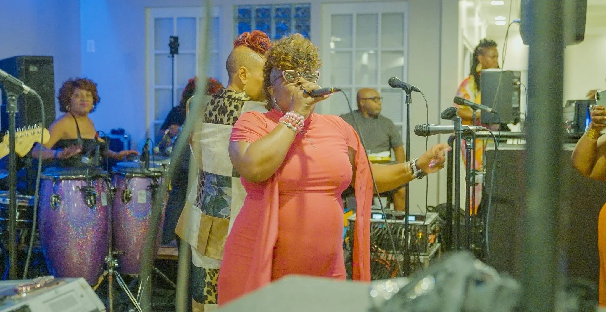 A woman wearing glasses and coral-colored dress singing into a microphone. Other people are in the background, including another woman with a colorful, patterned blouse and a person with pink glittery conga drums. The scene appears to be a lively ind