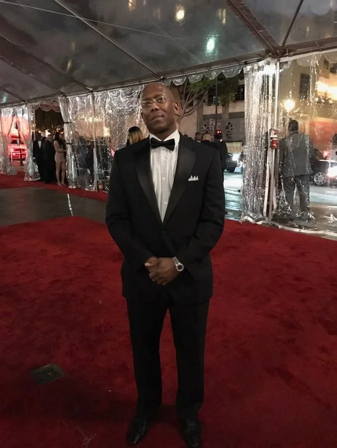 Man in a black tuxedo with a bow tie, standing on a red carpet at an event under a decorated tent.