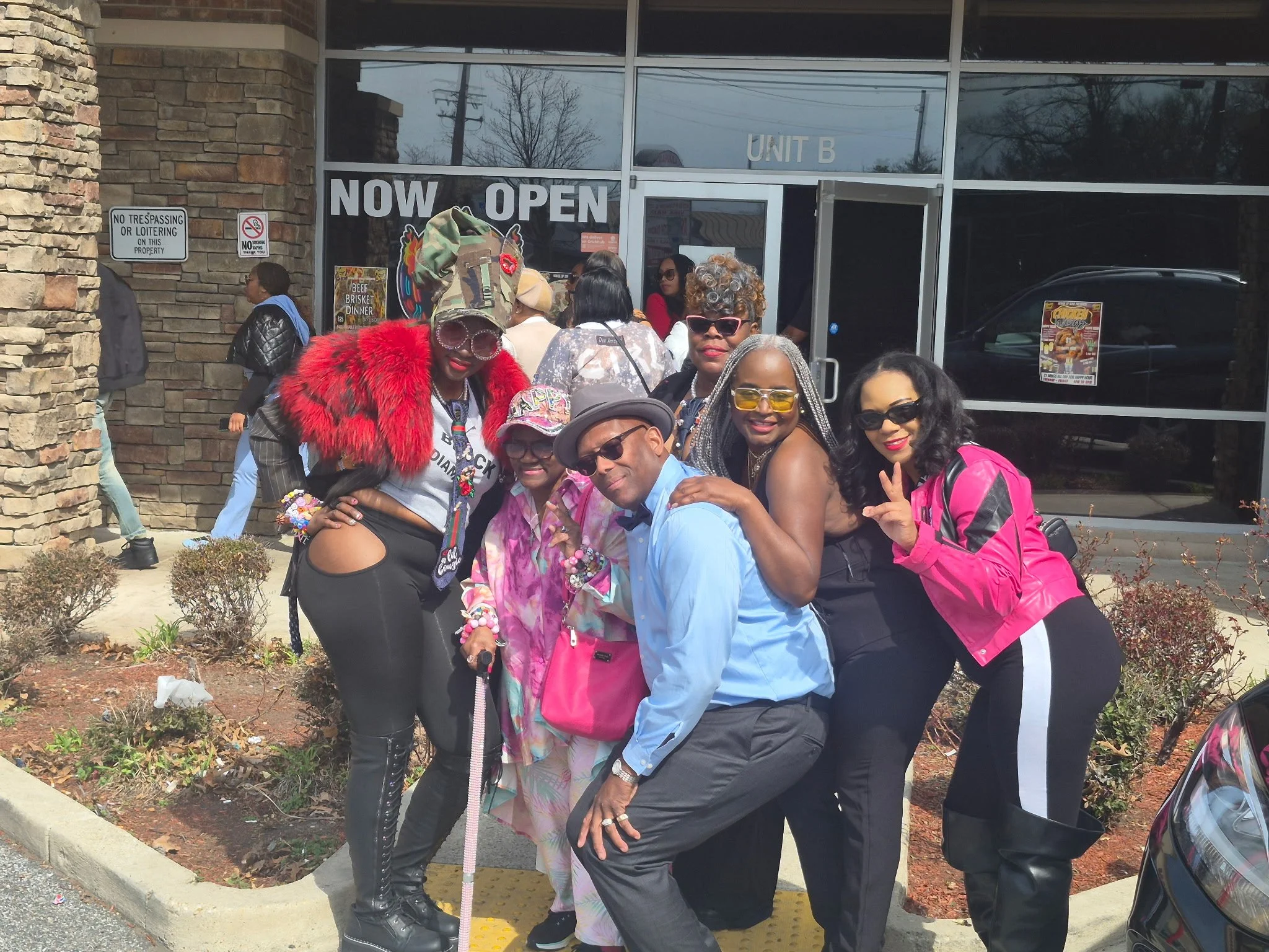 A group of diverse people posing for a photo outside of a building with a sign that says "NOW OPEN." The building has a brick and glass exterior, with some people wearing sunglasses, colorful clothing, and accessories. There are signs on the wall, in