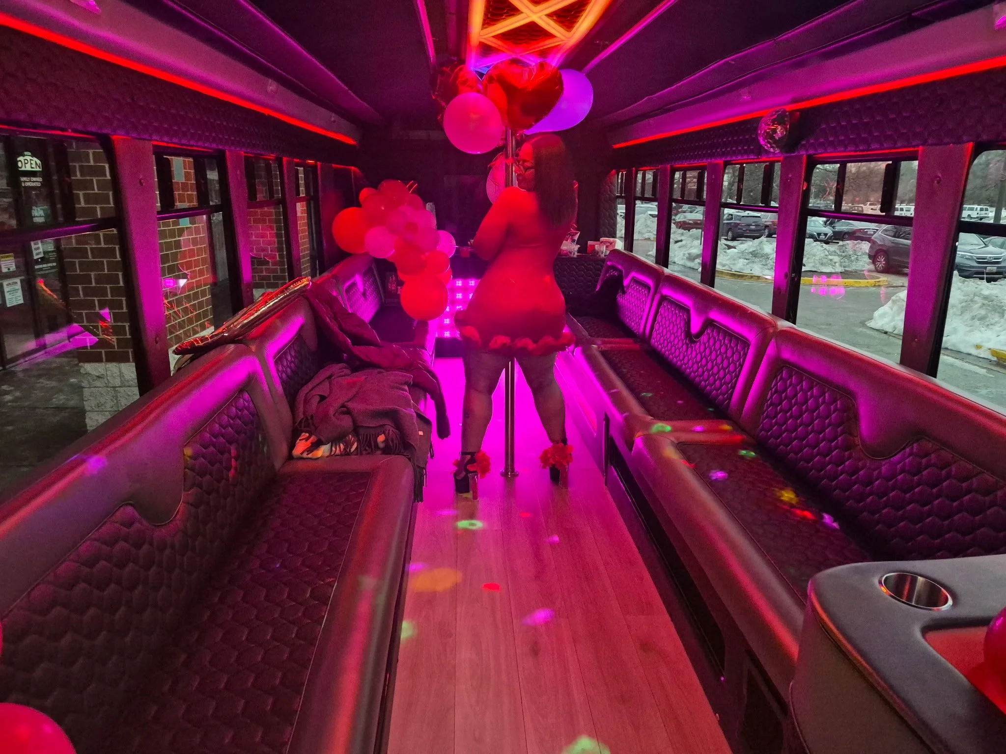 A woman in a dress holding a bunch of balloons stands inside a pink-lit party bus with patterned seats. Outside windows show a snowy parking lot.