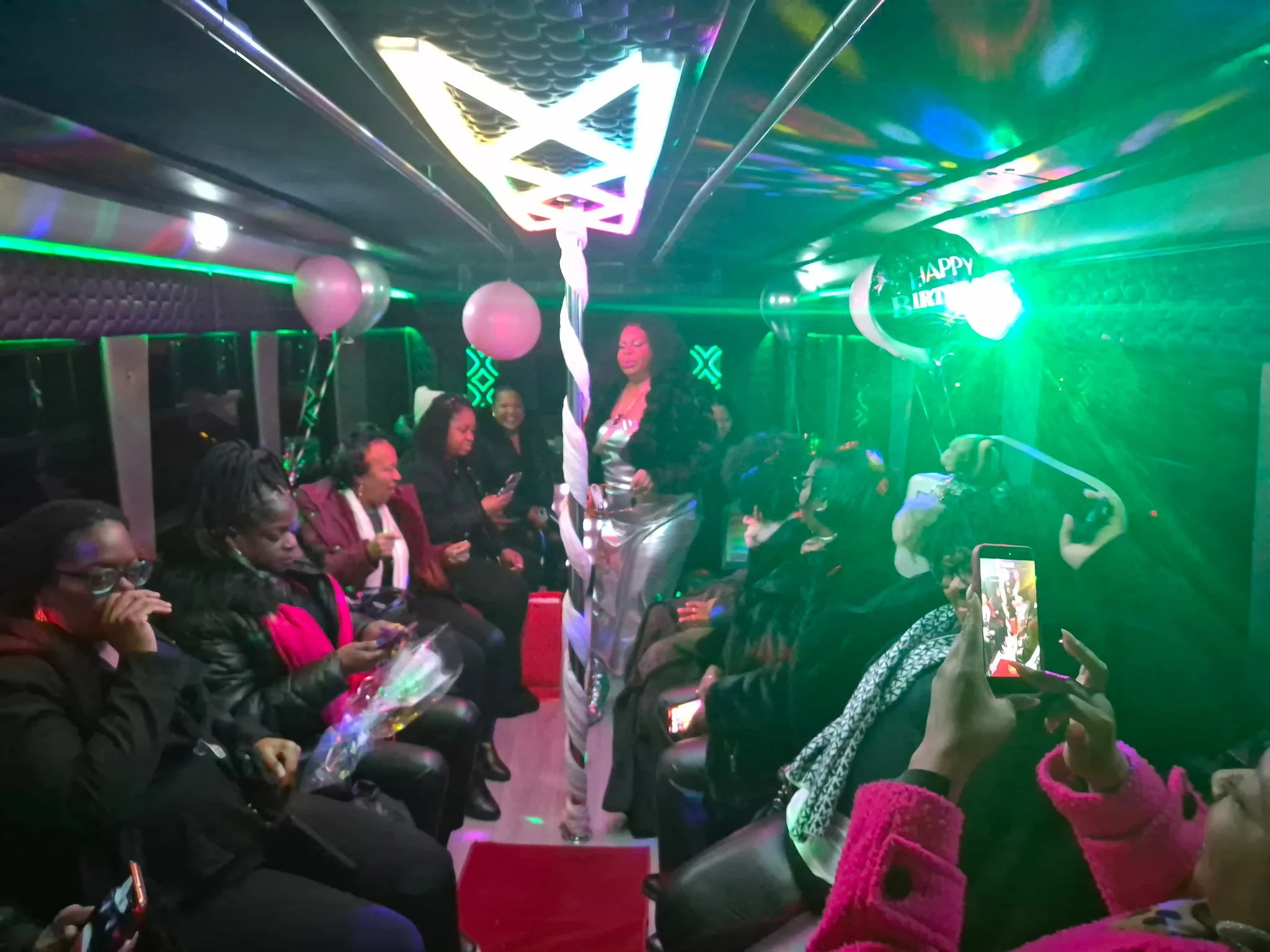 Indoor celebration with people seated, balloons, and green lighting, with a woman standing in the center near a decorative pole.