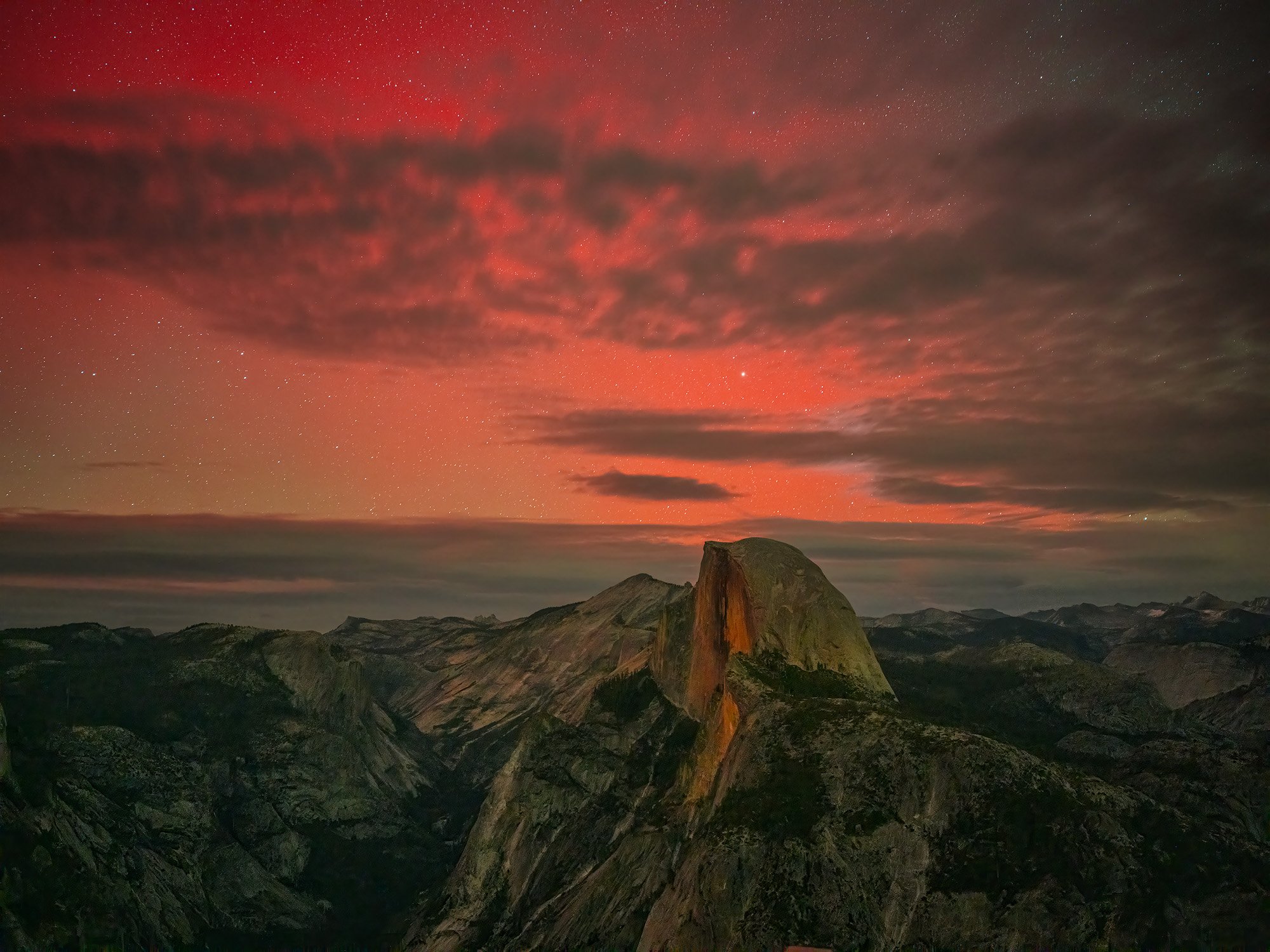 Nighttime view of Yosemite National Park with Half Dome, dark clouds, and a starry sky with a faint reddish hue by Charlotte Gibb.