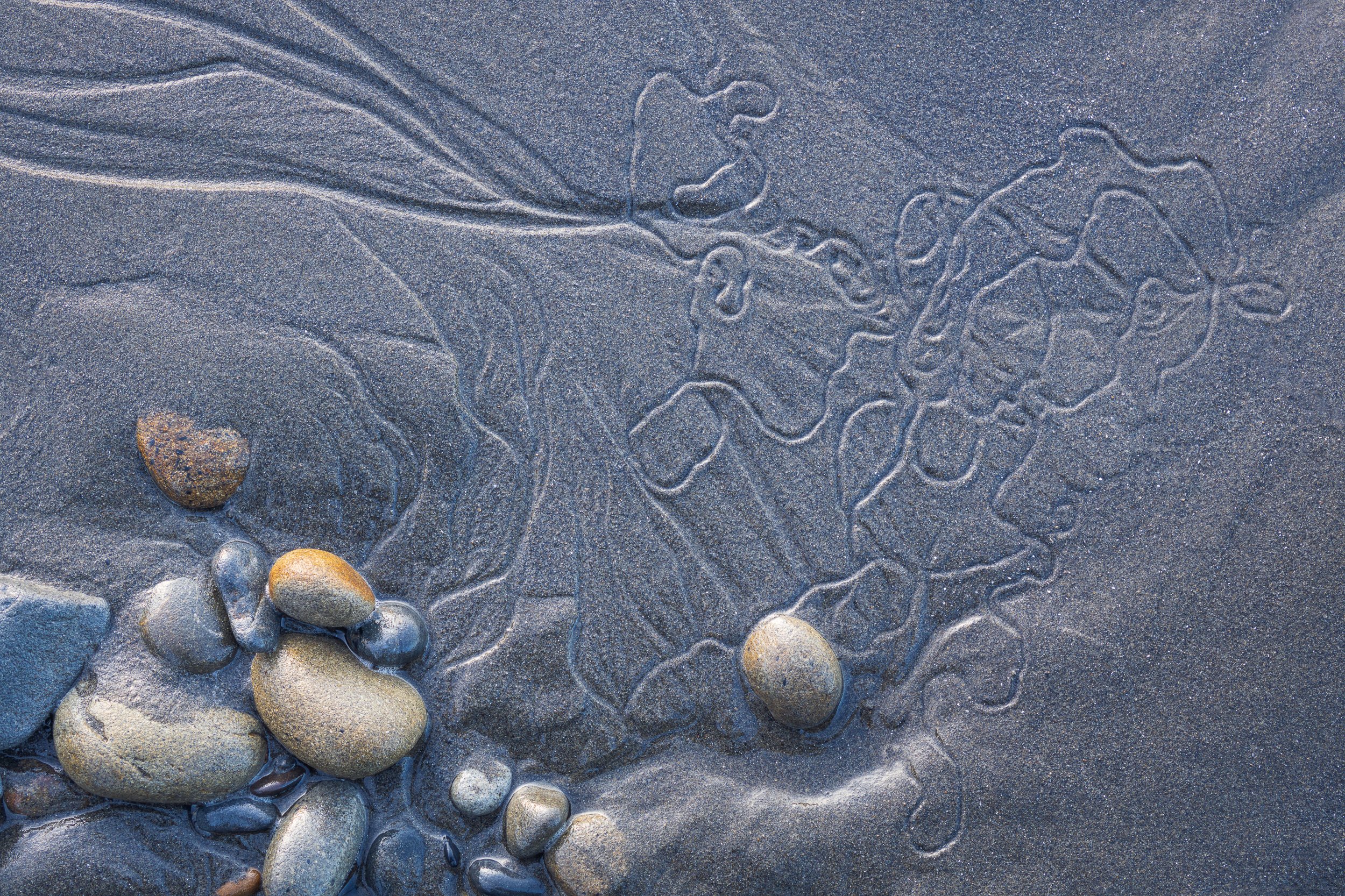 Rocks on a sandy beach with wind-created patterns and a faint heart shape in the sand by Sarah Marino.