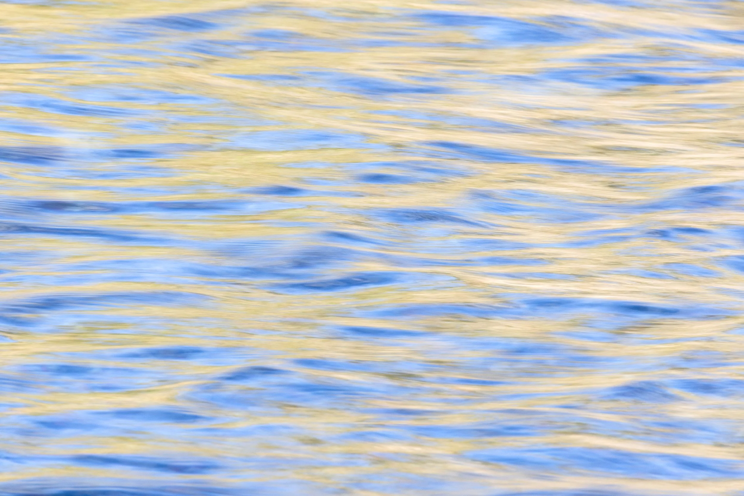 Close-up of water with ripples and reflections, showing blue and yellowish hues by Anna Morgan.