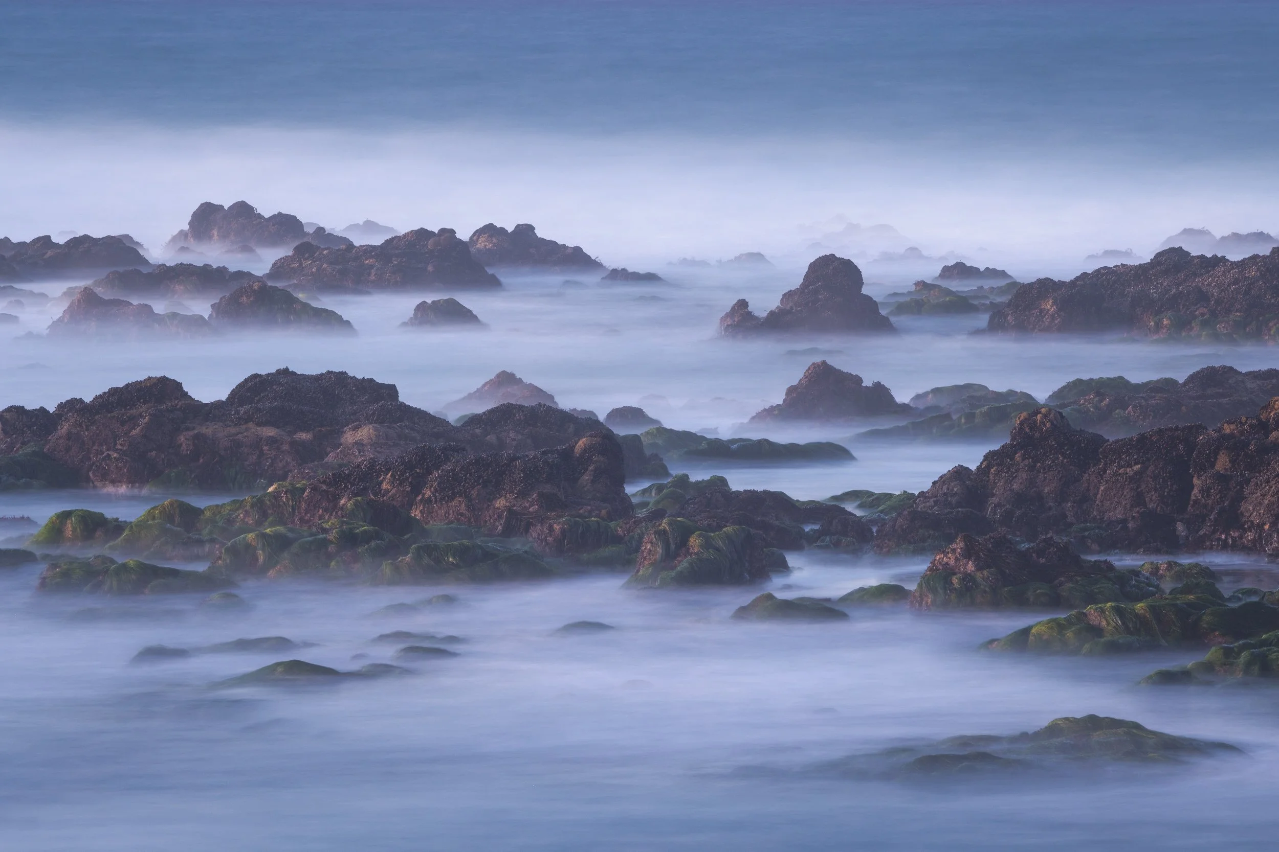 Rocky shoreline with moss-covered rocks and sea foam, ocean waves softly blurred, under a cloudy sky by Sarah Marino.