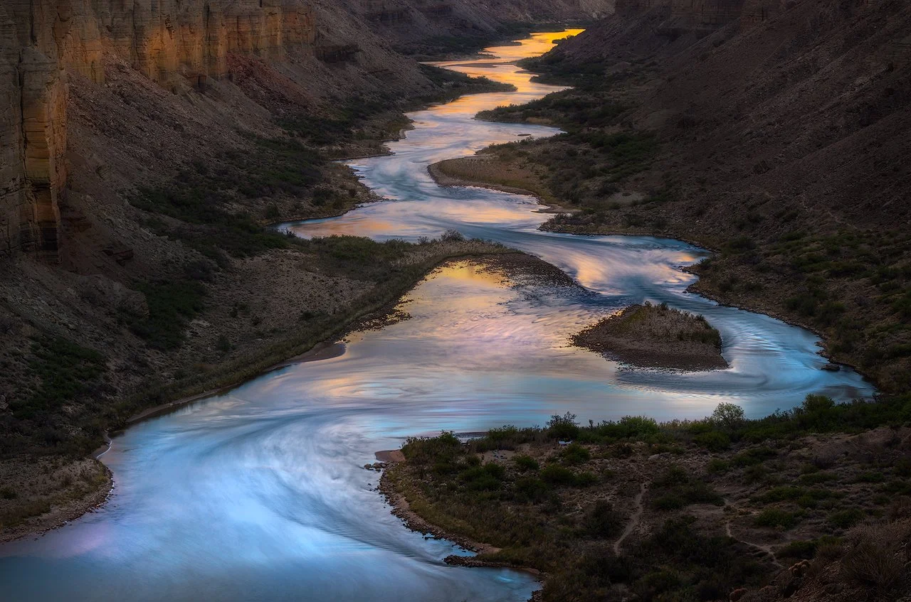 A winding river flowing through a canyon at sunset, with steep rocky cliffs on each side and sparse vegetation along the riverbanks by Charlotte Gibb.