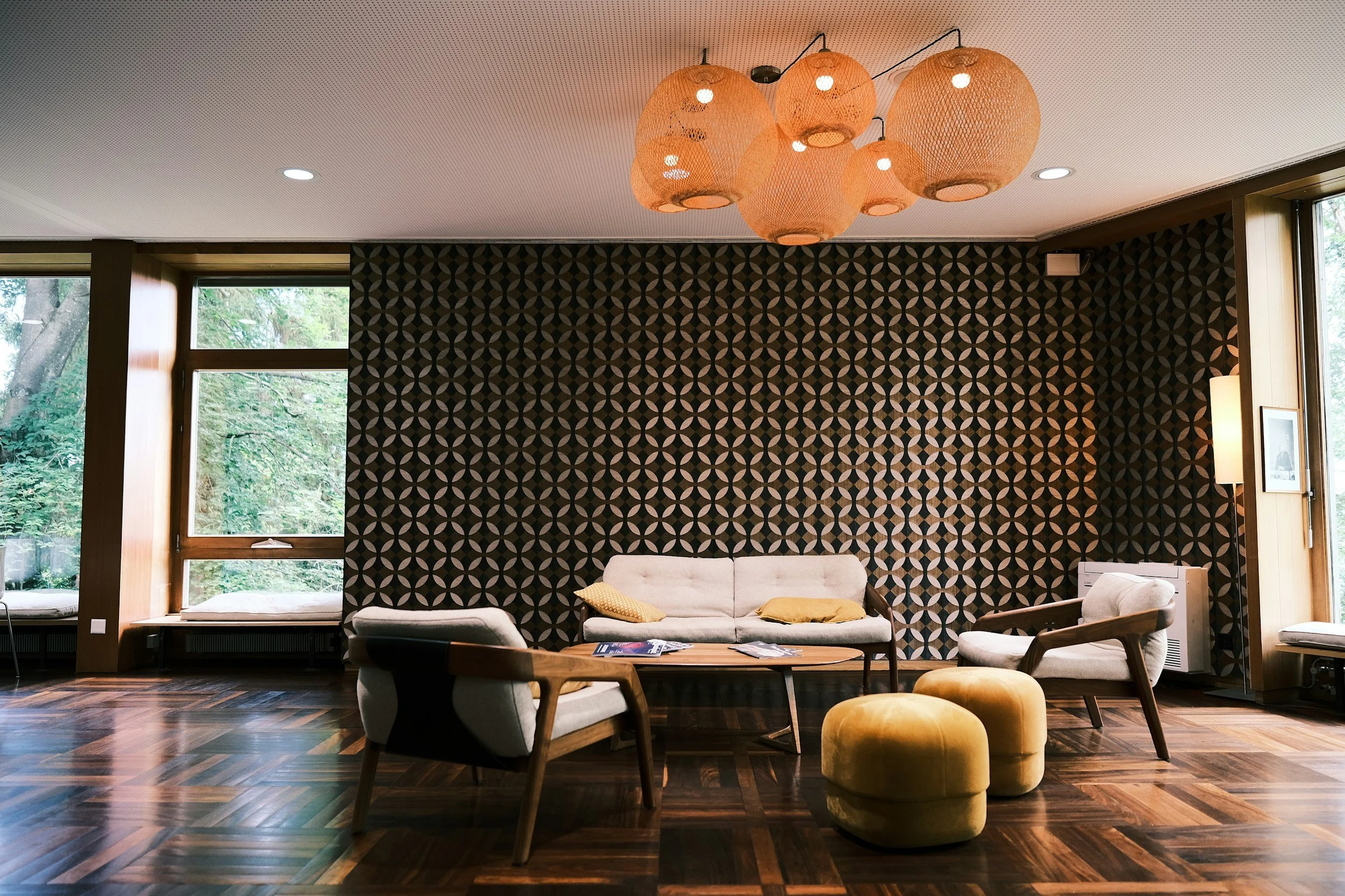 Modern living room with large windows, patterned black and beige wallpaper, beige sofa with pillows, wooden coffee table with magazines, and a floral chandelier lighting fixture.