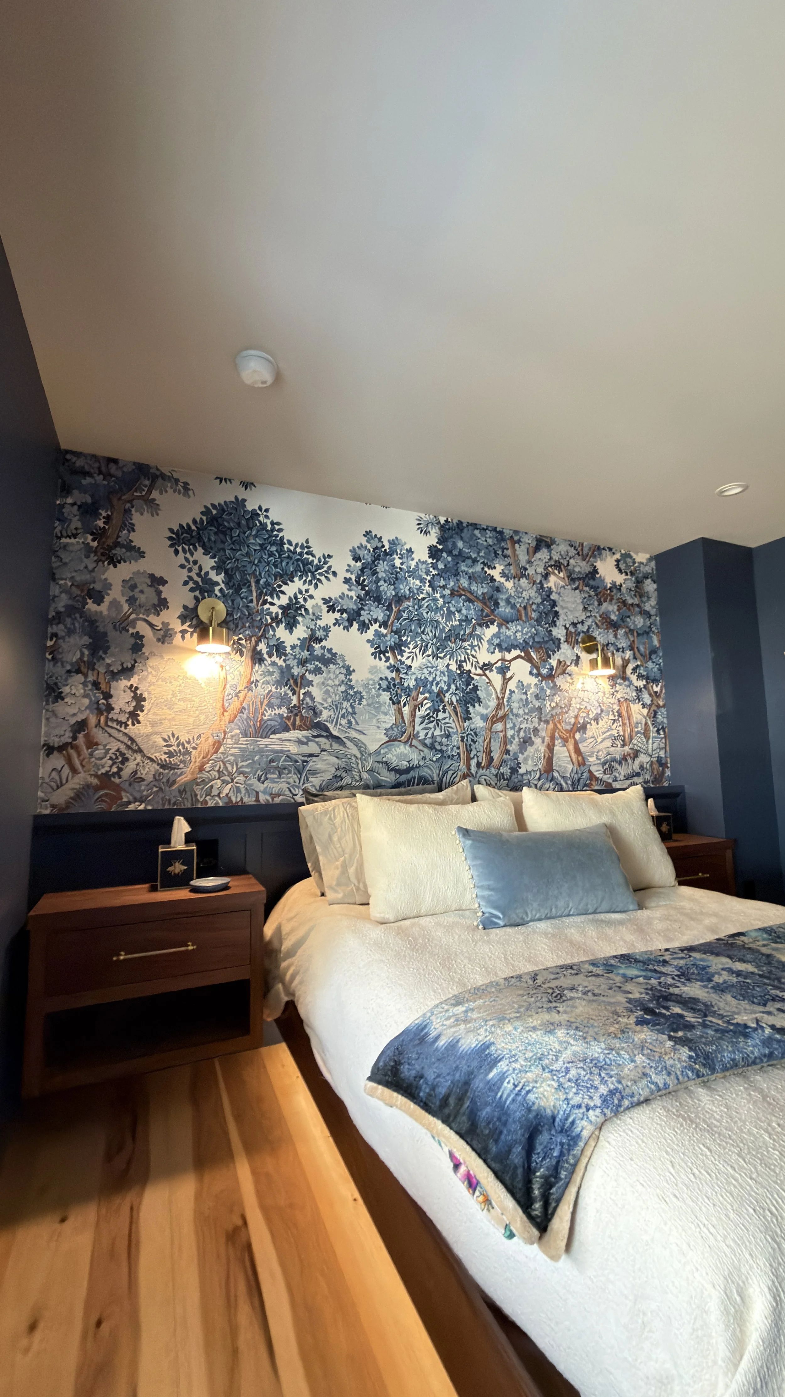 A bedroom with a bed featuring cream-colored bedding and blue accent pillows. Behind the bed is a decorative blue mural of trees, accompanied by wall-mounted lamps with gold accents. There are wooden nightstands on each side of the bed and a ceiling smoke detector.