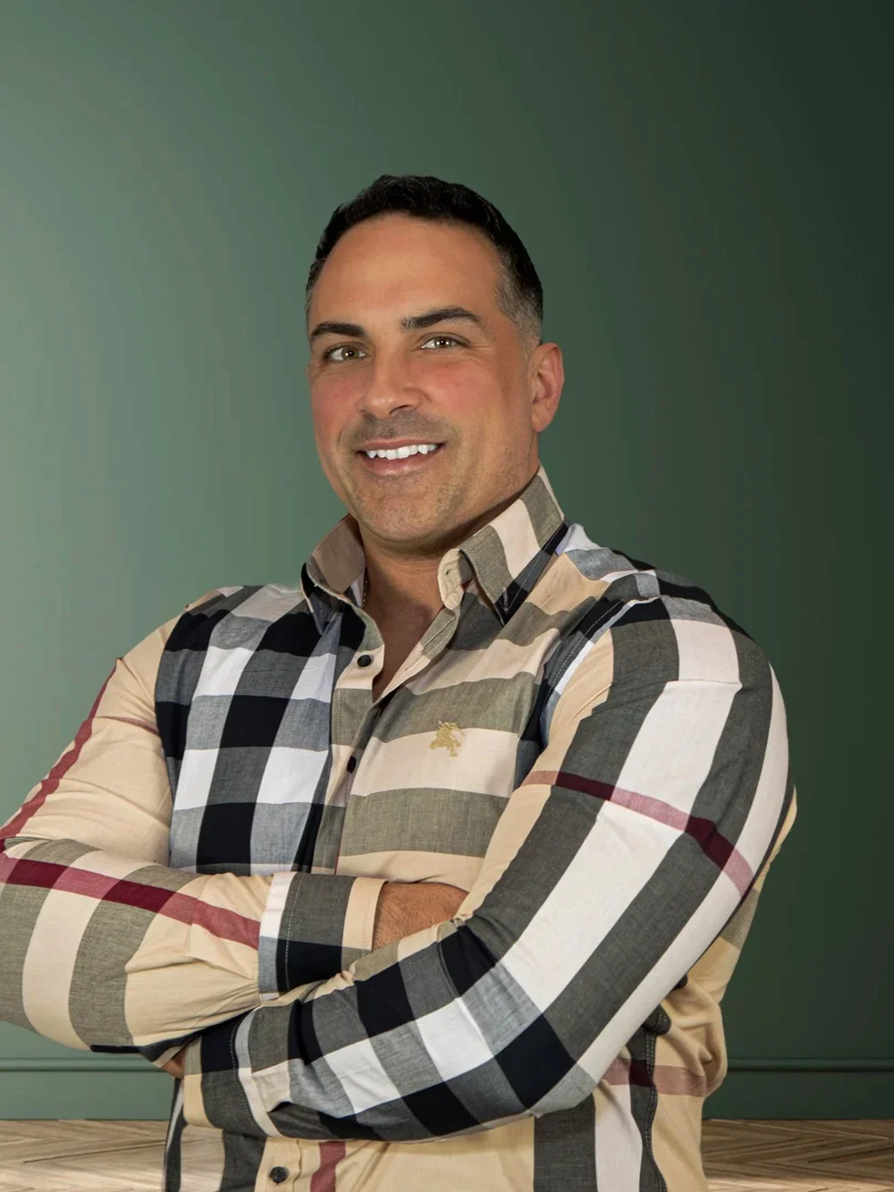 A smiling man with crossed arms wearing a plaid shirt standing against a gradient green background.