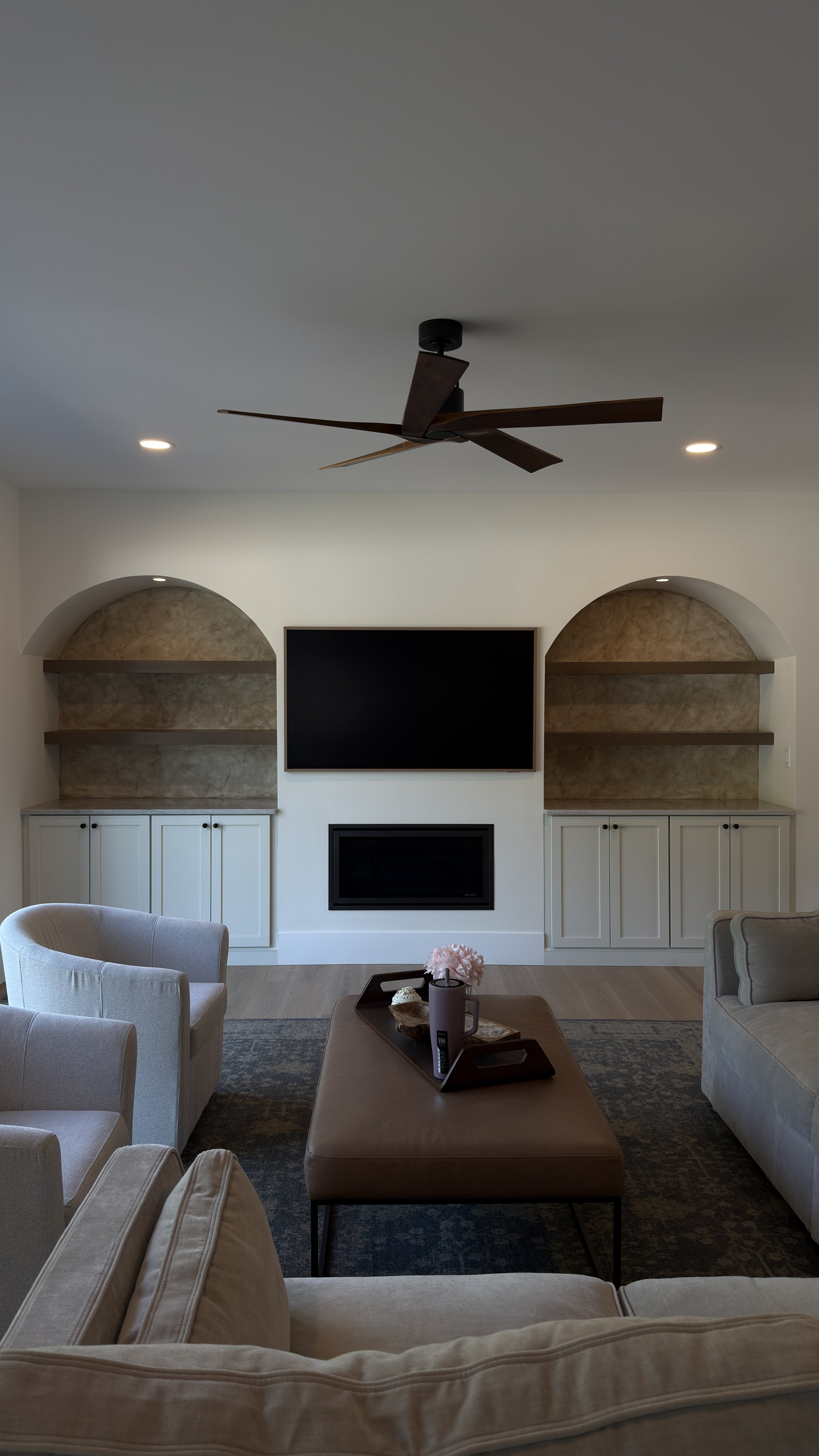 Living room with beige sofas, a large flat-screen TV above a fireplace, built-in shelves on either side, a dark brown ottoman with a tray and decorative objects, and a ceiling fan.