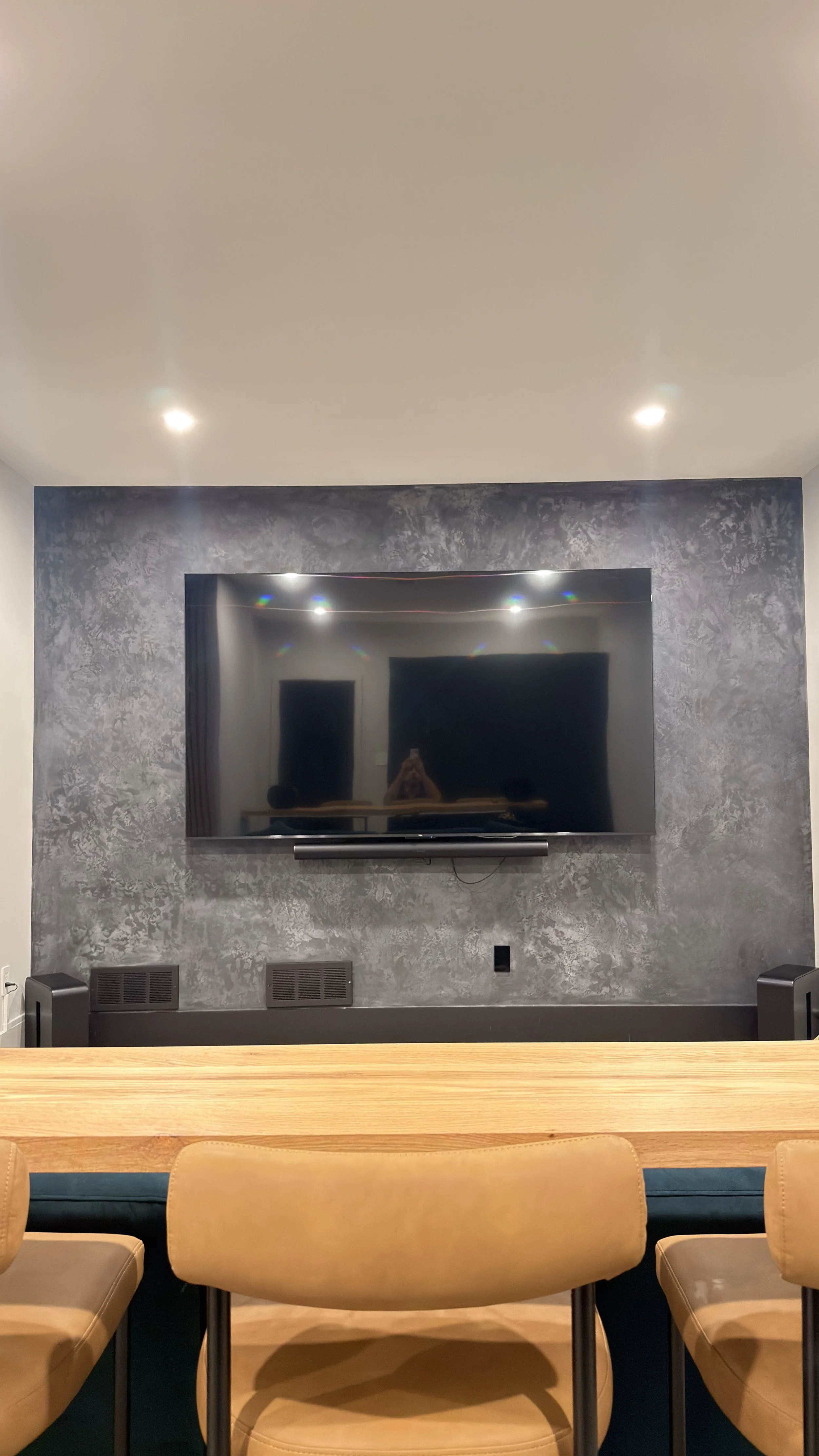 Living room with a flat-screen TV mounted on a textured dark gray wall, beige chairs in the foreground, and a wooden table.