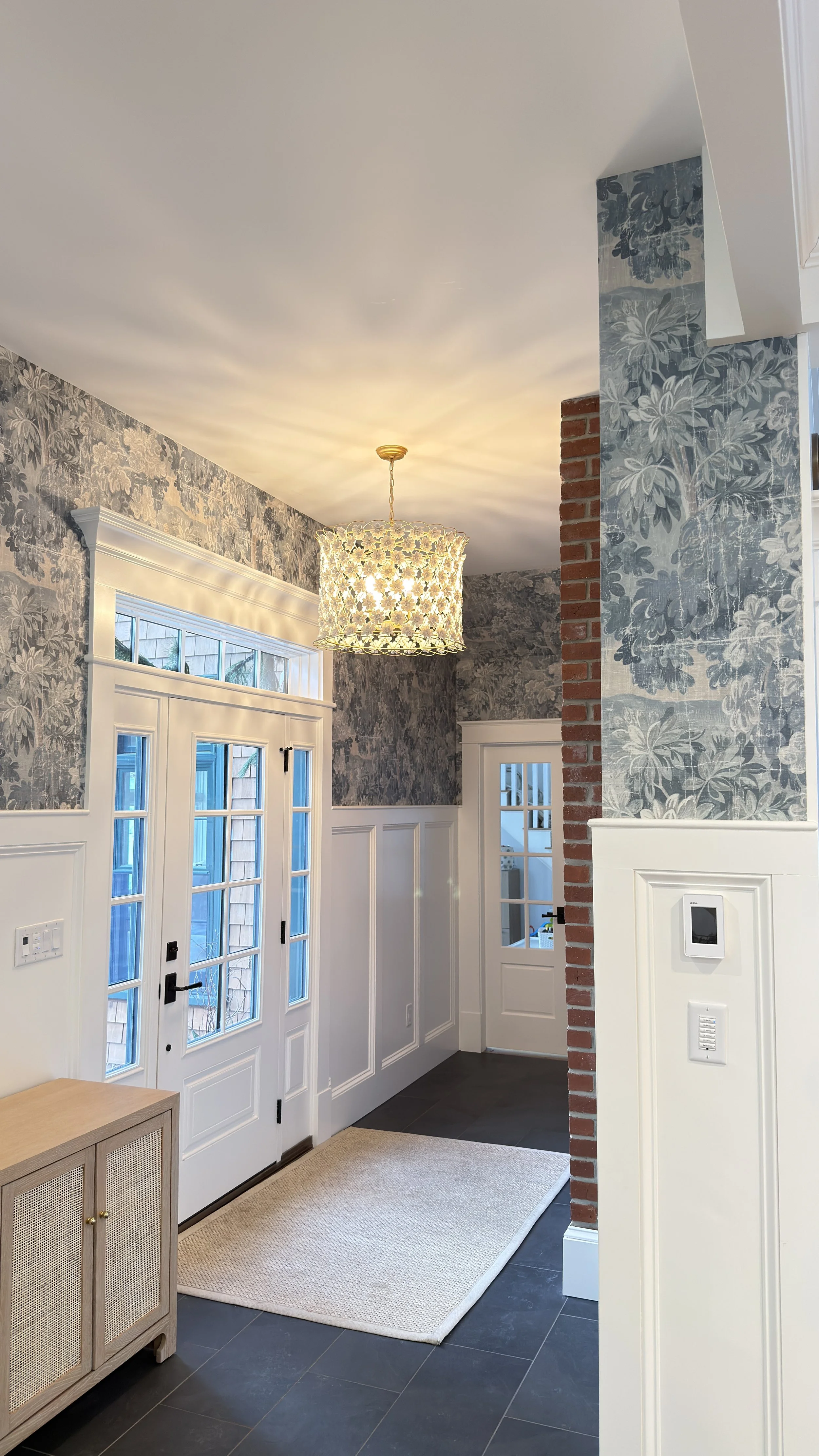 Entryway with white door and glass panels, floral wallpaper, chandelier, small furniture, and a rug.