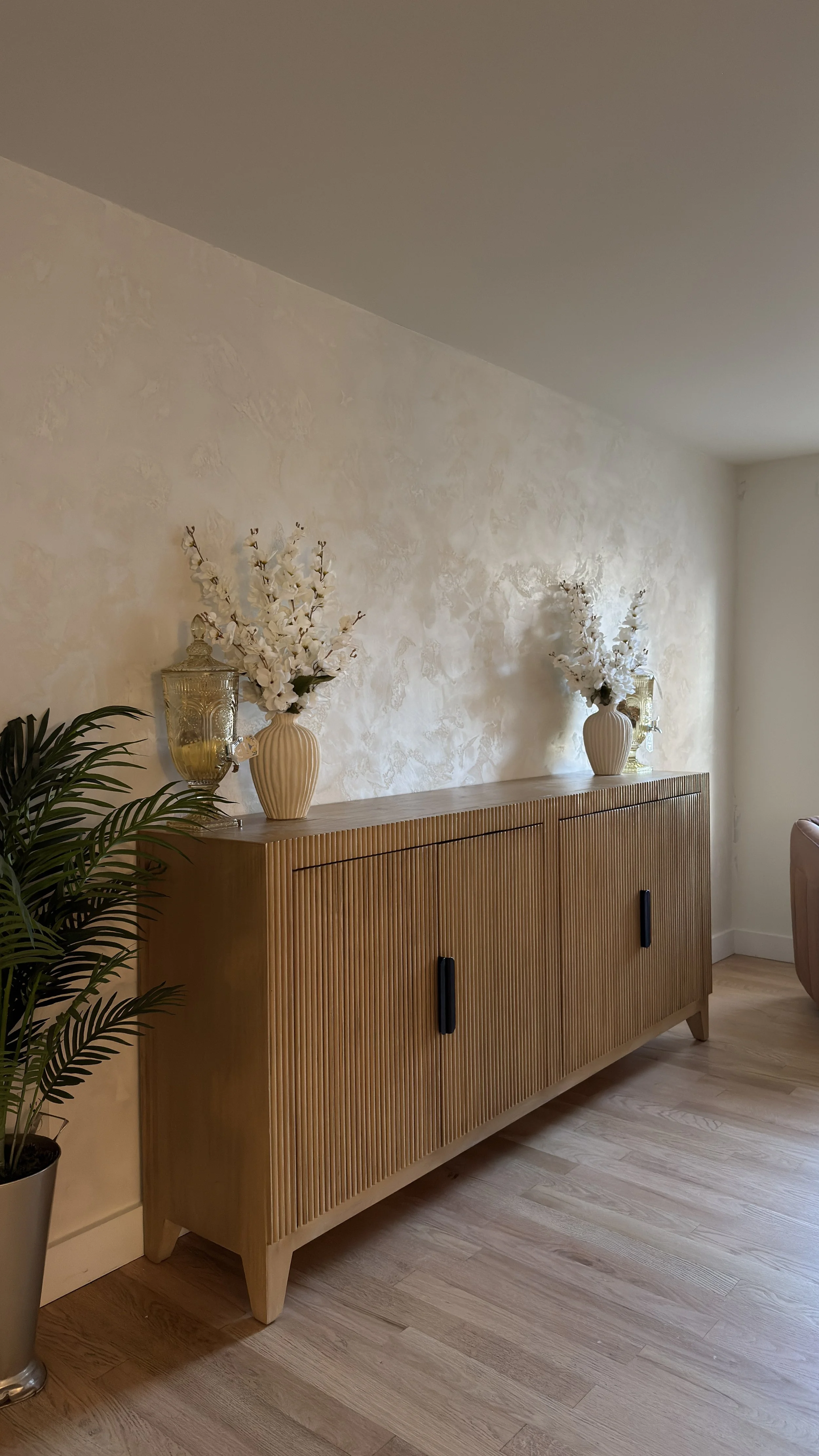 A wooden sideboard with vertical grooves, decorated with two white vases filled with white flowers, a glass jar, and a green plant nearby. The wall behind has a textured, light-colored finish, and the floor is wood with a light finish.