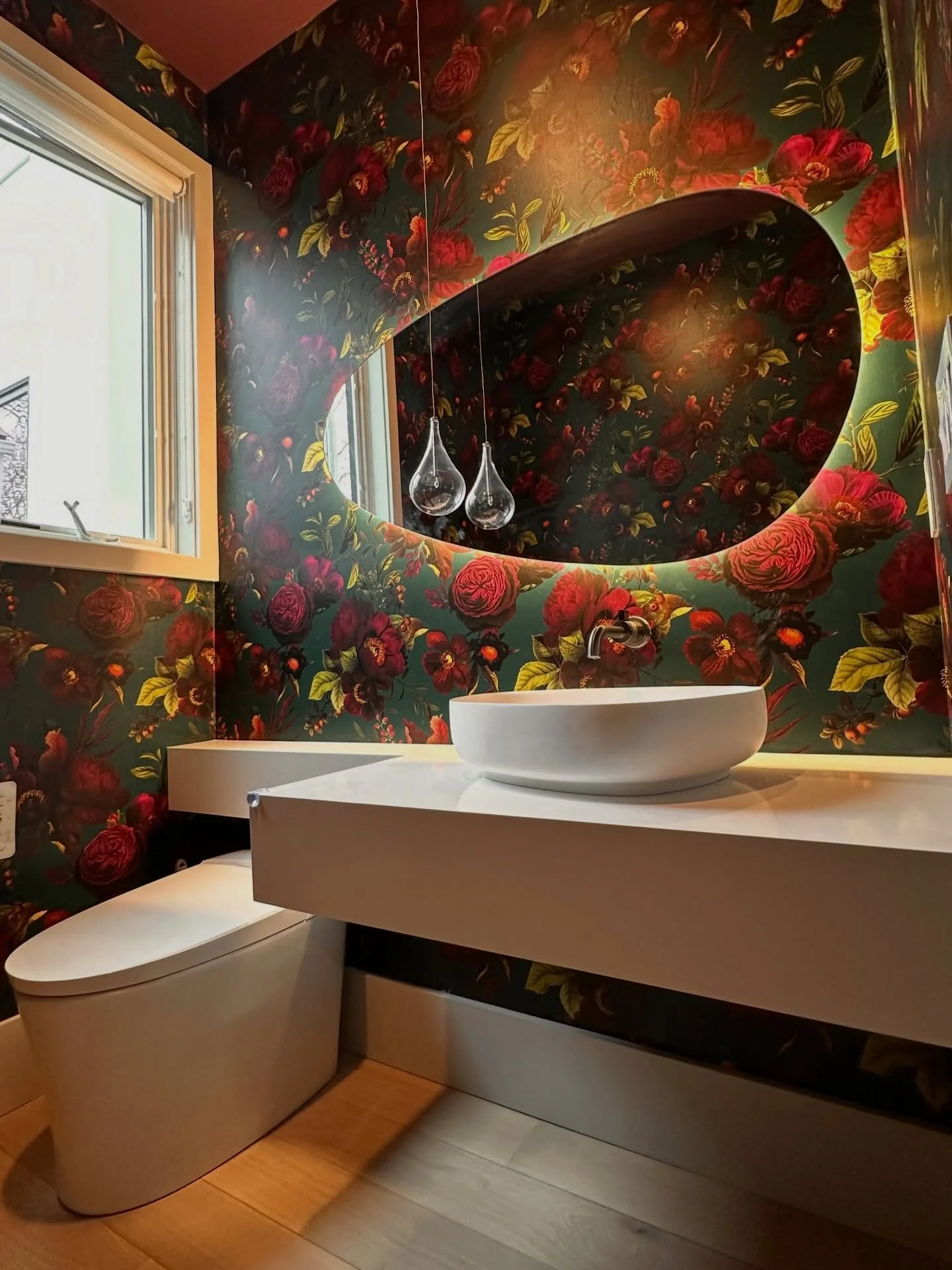 A bathroom with vibrant floral wallpaper, a modern white toilet, a white vanity with a vessel sink, and a mirror with two hanging pendant lights.