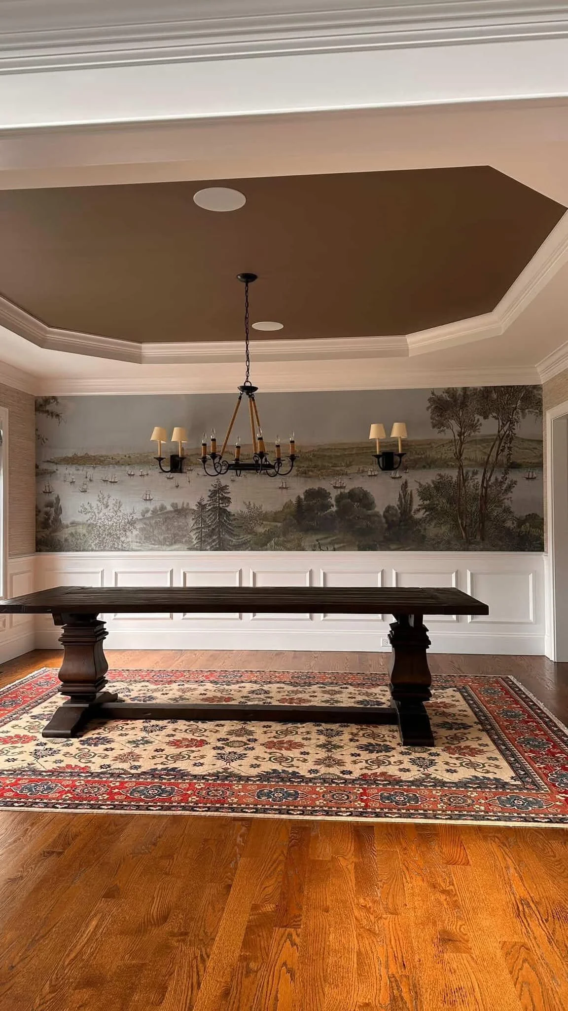 Room with hardwood floor, a patterned area rug, a long dark wooden table, wainscoting, a mural of a landscape with trees, water, and boats, and a chandelier hanging from the ceiling.