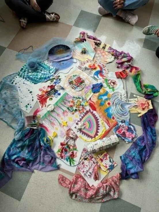 Colorful artwork and craft supplies arranged on the floor, surrounded by children’s legs and shoes.