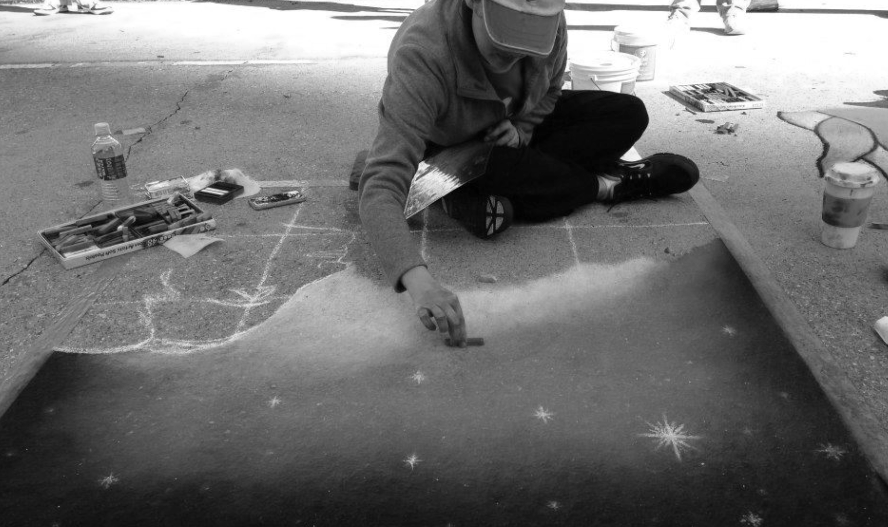 A person sitting on the ground creates chalk art on a sidewalk, surrounded by chalk, a water bottle, and beverages, with some of the artwork resembling snowflakes and star shapes.