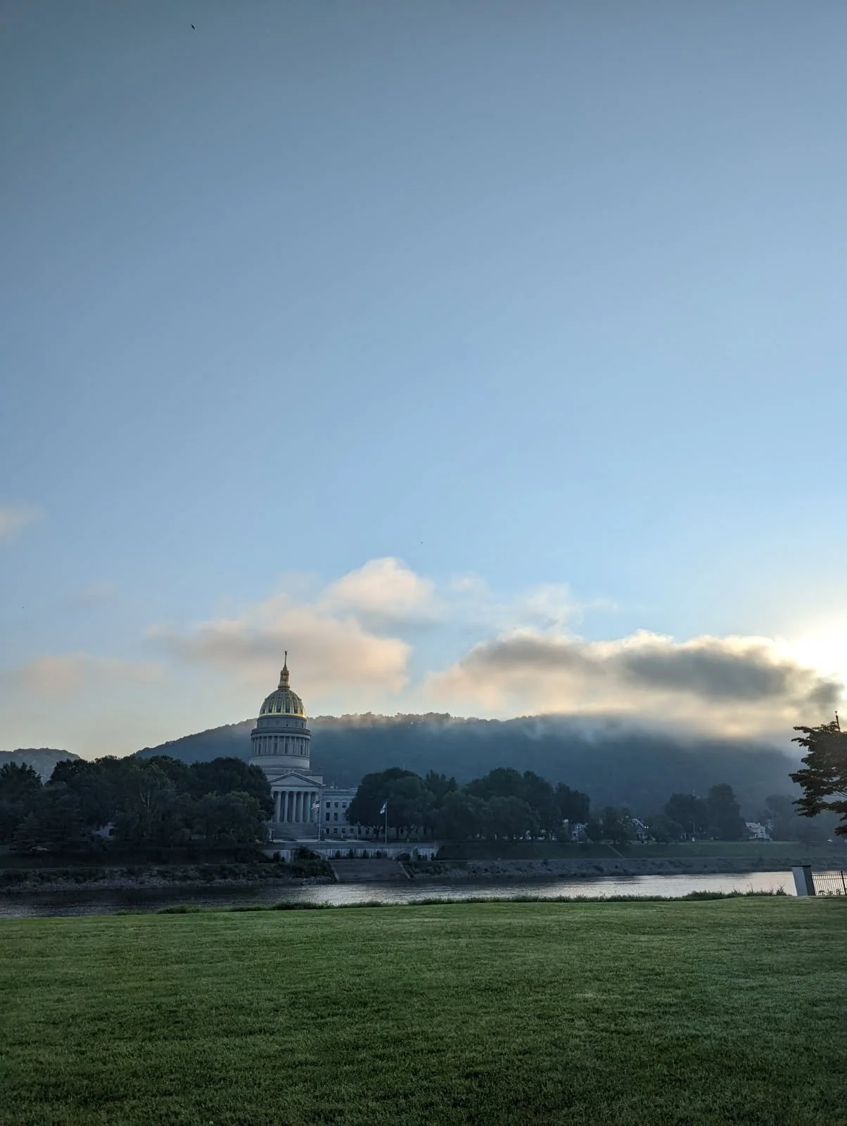 The image shows the United States Capitol building with its iconic dome, situated behind a line of trees, across a body of water, under a sky with scattered clouds and early morning sunlight. In the foreground, there is a well-manicured grassy area.