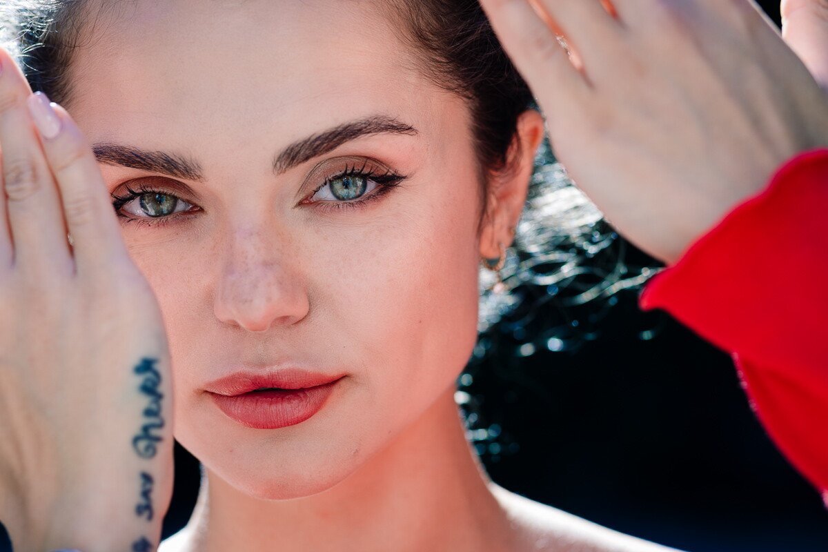 A close-up portrait of a woman with blue eyes, freckles, and light makeup, holding her hands near her face with fingers spread, with another person partially visible in a red sleeve touching her hair.