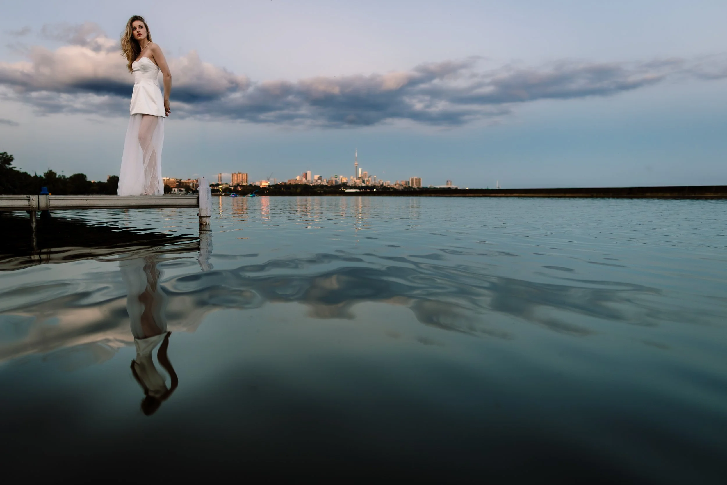 A woman in a white dress standing on a dock by the water with a city skyline, including the CN Tower, visible in the distance under a partly cloudy sky.