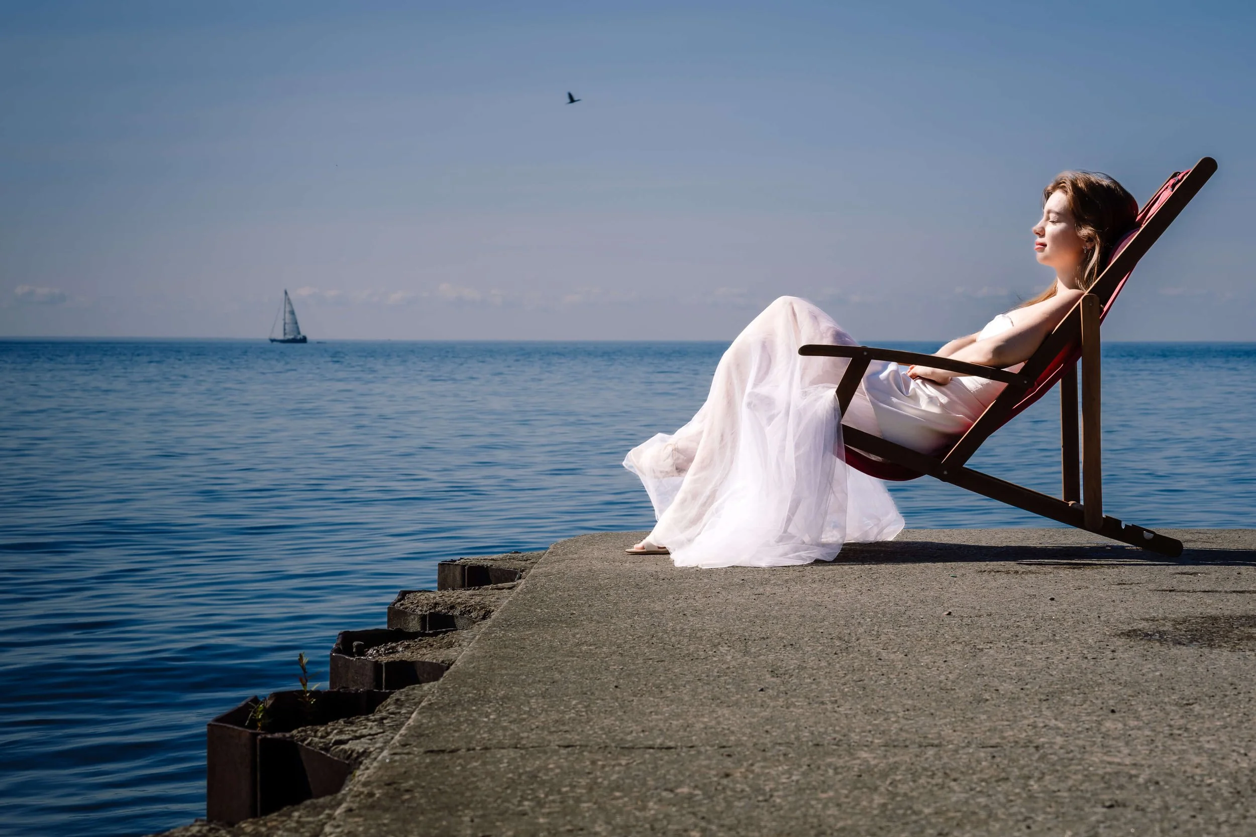 A woman in a white dress relaxing on a lounge chair by the water on a concrete pier, with a sailboat and bird in the sky in the background.