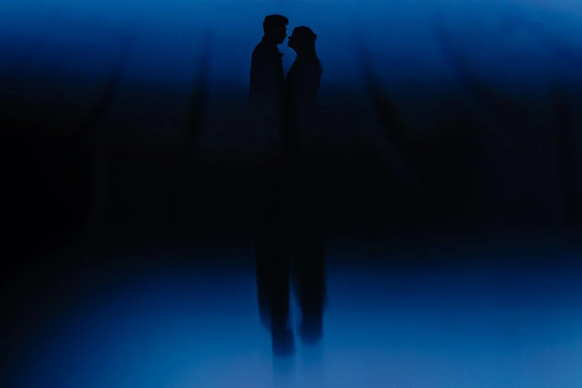 Silhouette of a couple facing each other, standing close against a blue sky, with their reflection visible on a dark shiny surface.