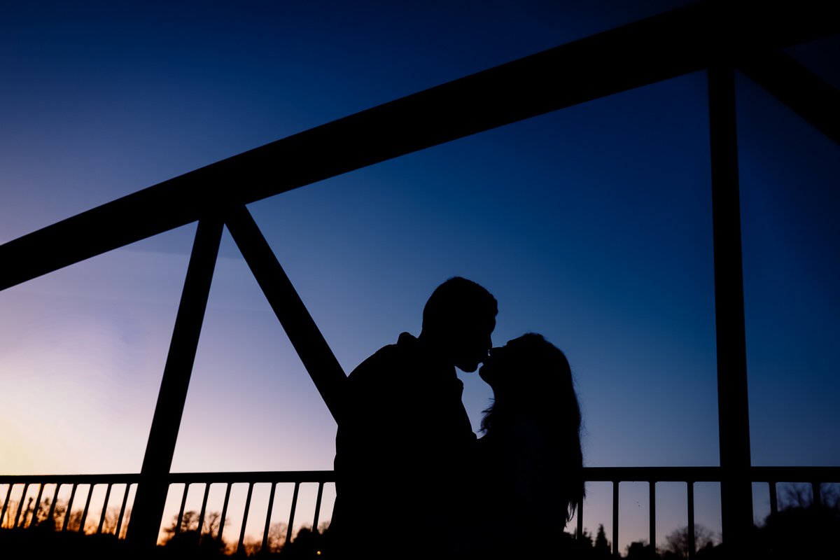 Silhouetted couple sharing a kiss or intimate moment on a bridge at sunset or dusk.