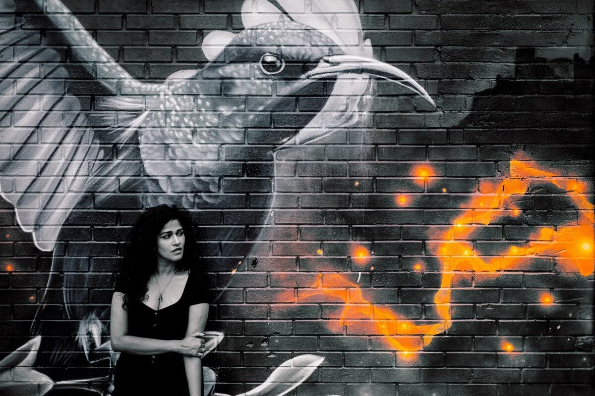 A woman with curly hair and a black shirt standing in front of a black and white street art mural of a bird on a brick wall, with orange glowing elements.