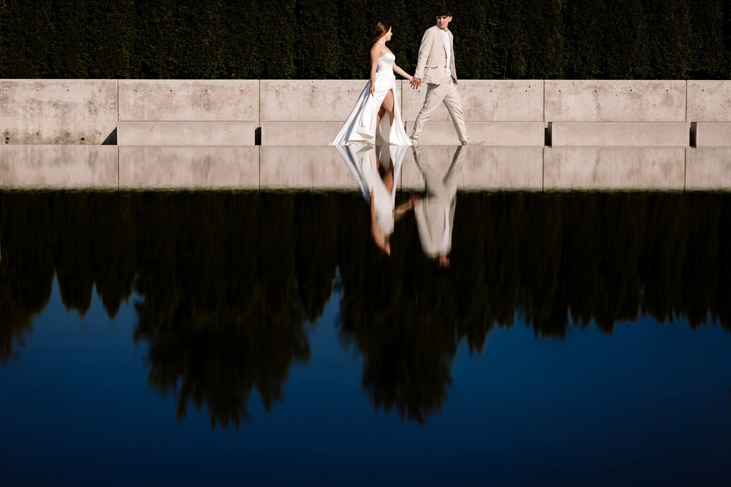 A couple in wedding attire walking hand in hand along a concrete wall beside a reflective body of water, with greenery in the background.