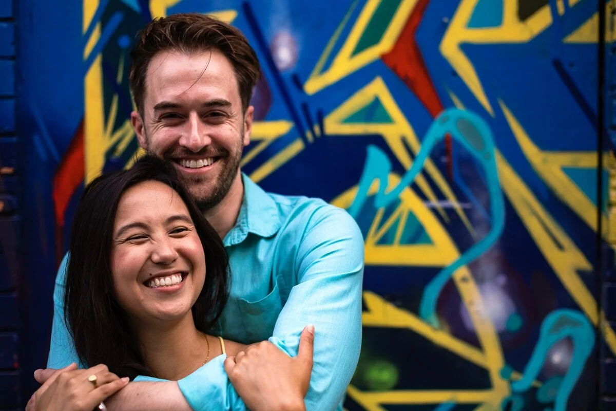 A smiling couple hugging in front of a graffiti-covered wall with bold blue, yellow, red, and teal abstract designs.