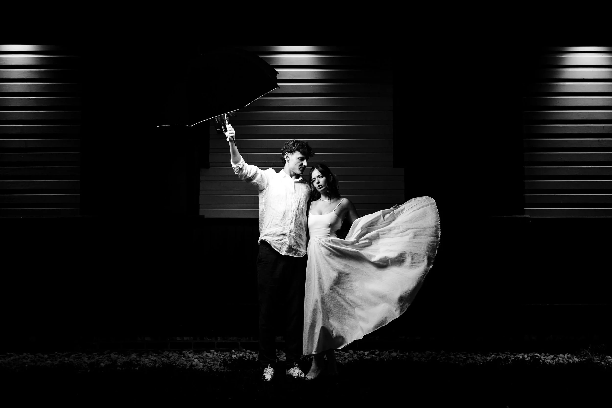 Black and white photo of a man and woman standing outdoors against a background of horizontal siding. The man is holding a large umbrella and looking at the woman, who is dressed in a flowing gown, and is holding the fabric of her dress.