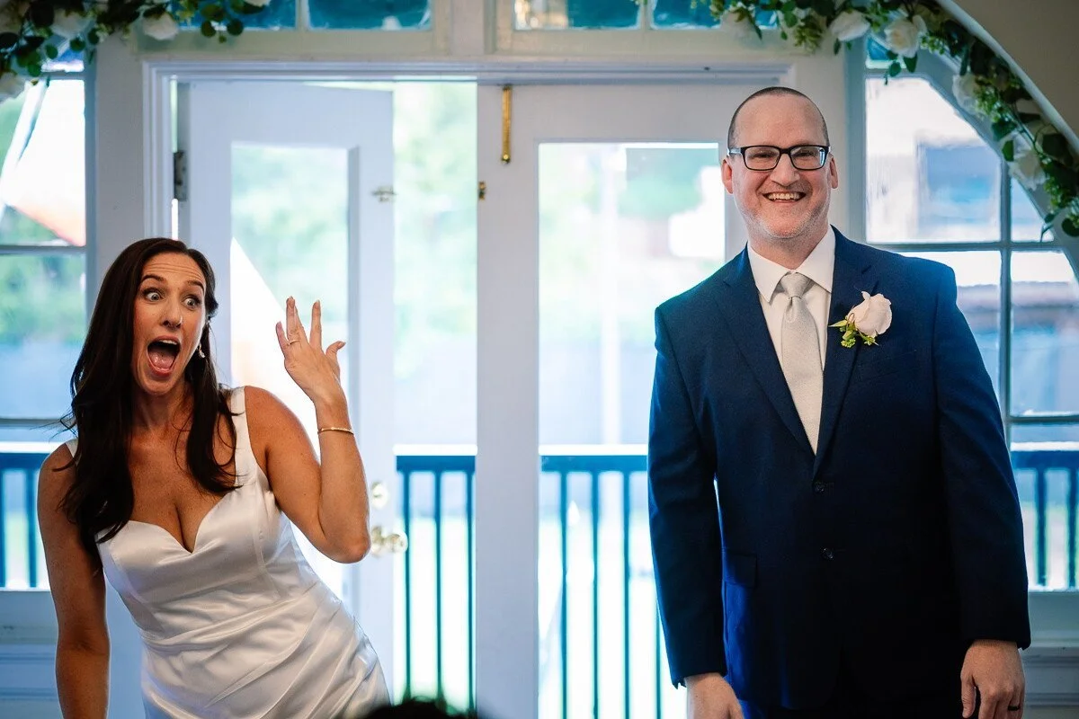 A woman in a white wedding dress with long dark hair and a man in a blue suit and glasses standing indoors with a floral arch overhead, at a wedding reception.