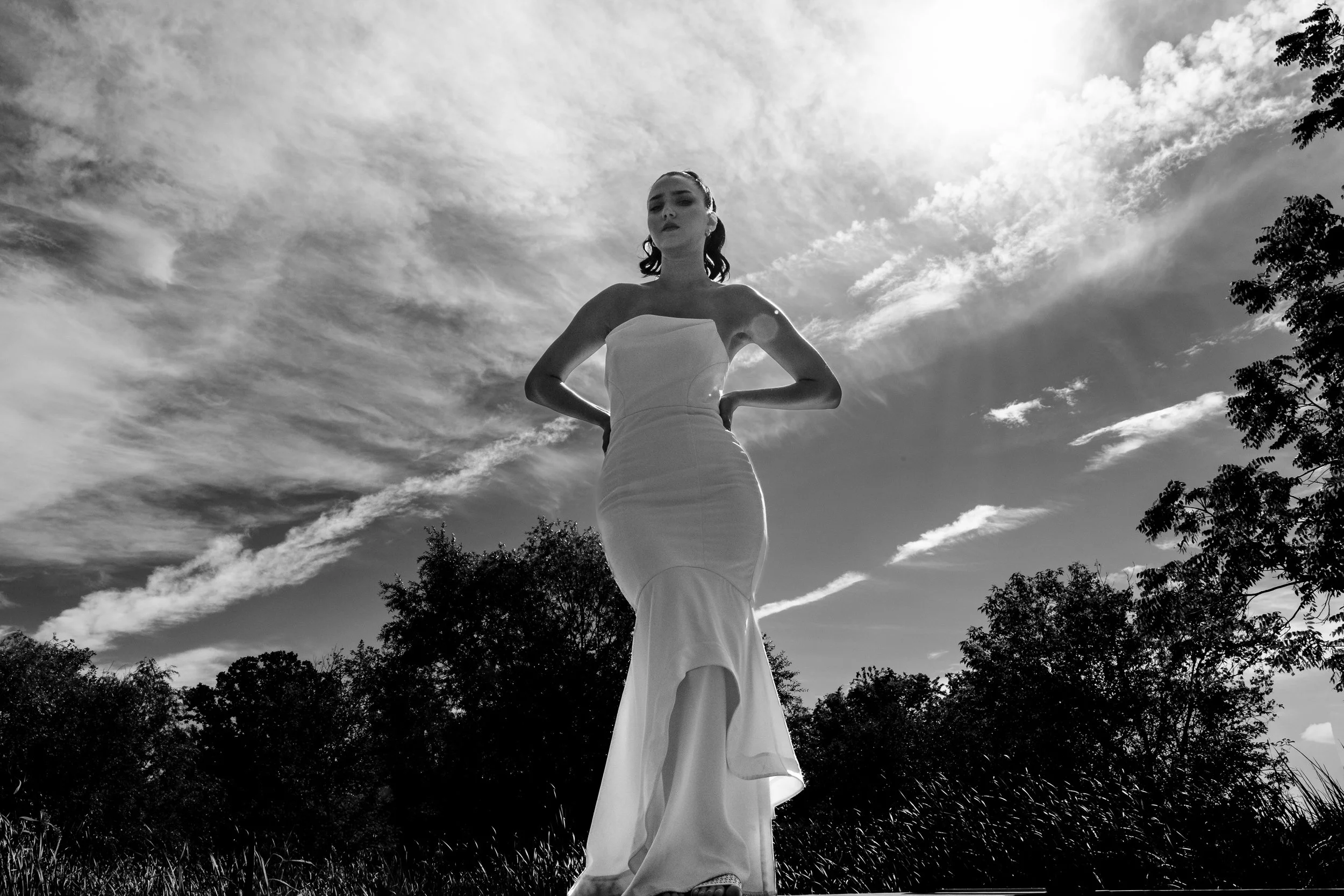 A woman standing outdoors in a strapless dress with her hands on her hips, looking solemn, with her hair in a ponytail. The photo is in black and white, and the sky is cloudy with trees at the bottom of the image.