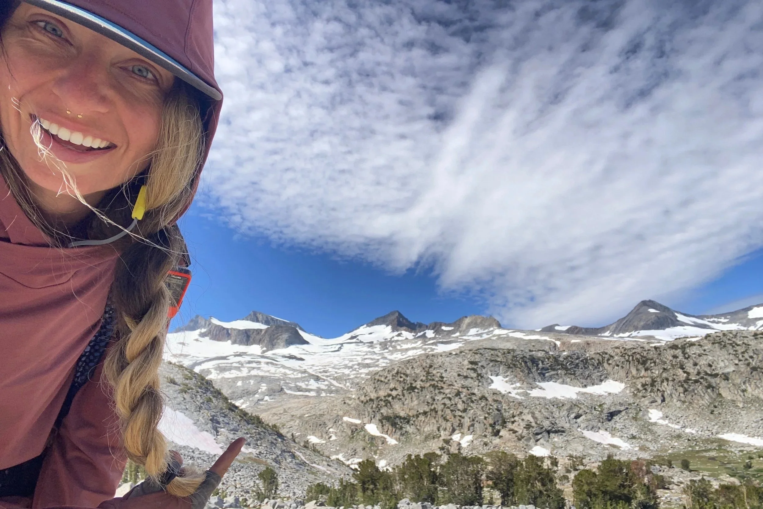 A woman with long blonde hair braided over her shoulder, wearing a pinkish waterproof jacket and a cap, smiling and pointing towards snow-capped mountains and cloudy sky in the background.