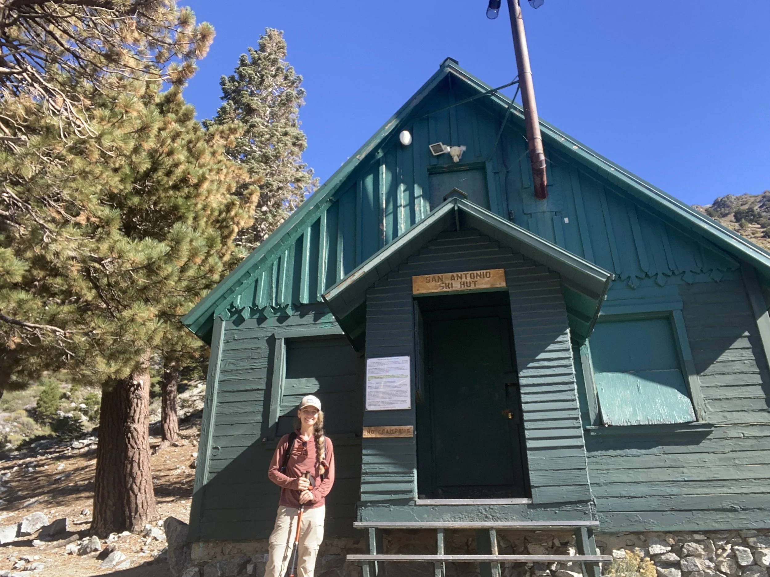 A woman standing outside a teal-colored wooden building labeled 'San Antonio Ski Hut' with trees and mountains in the background, under a clear blue sky.