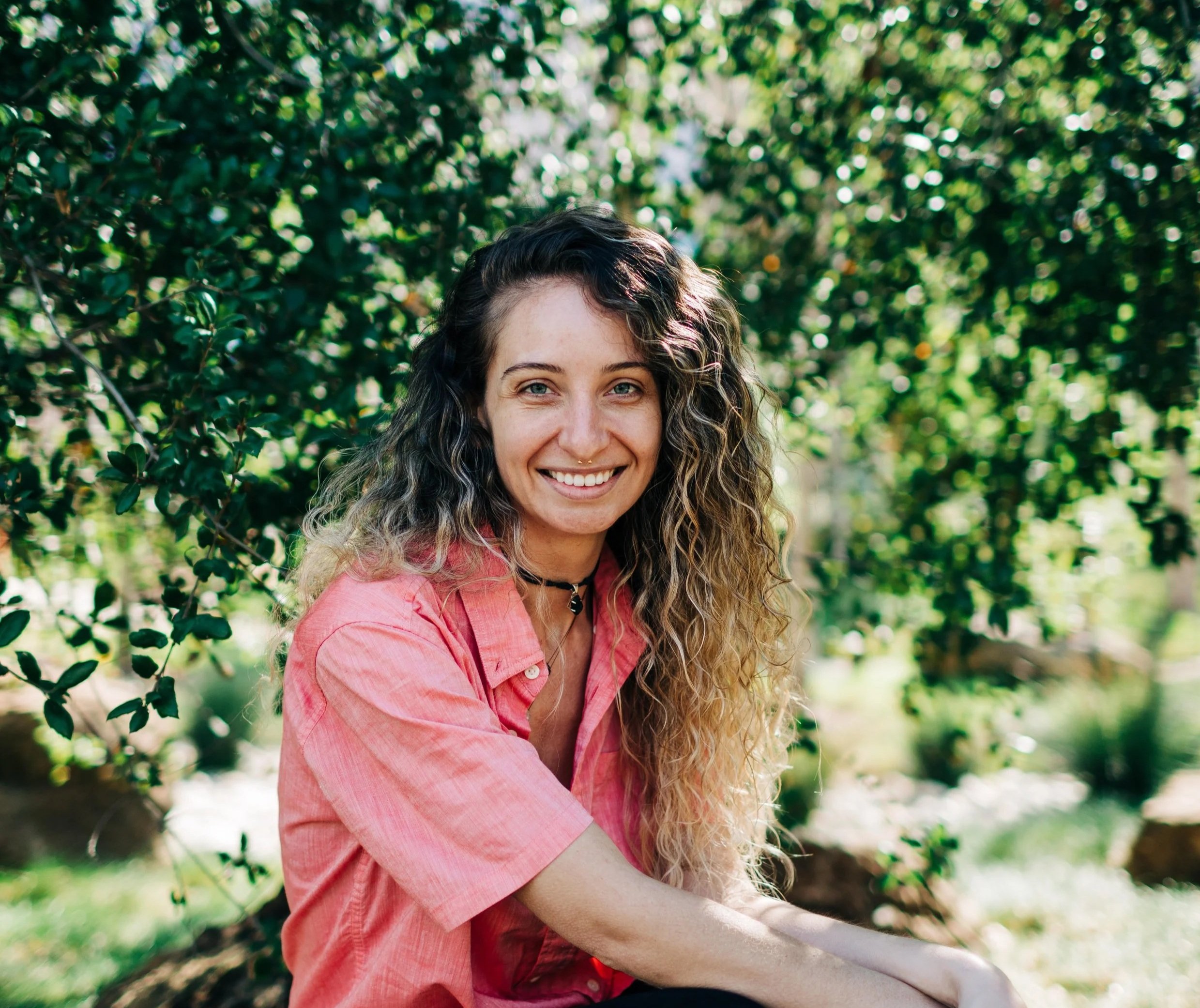 A woman with curly hair smiling outdoors among green foliage, wearing a pink shirt.