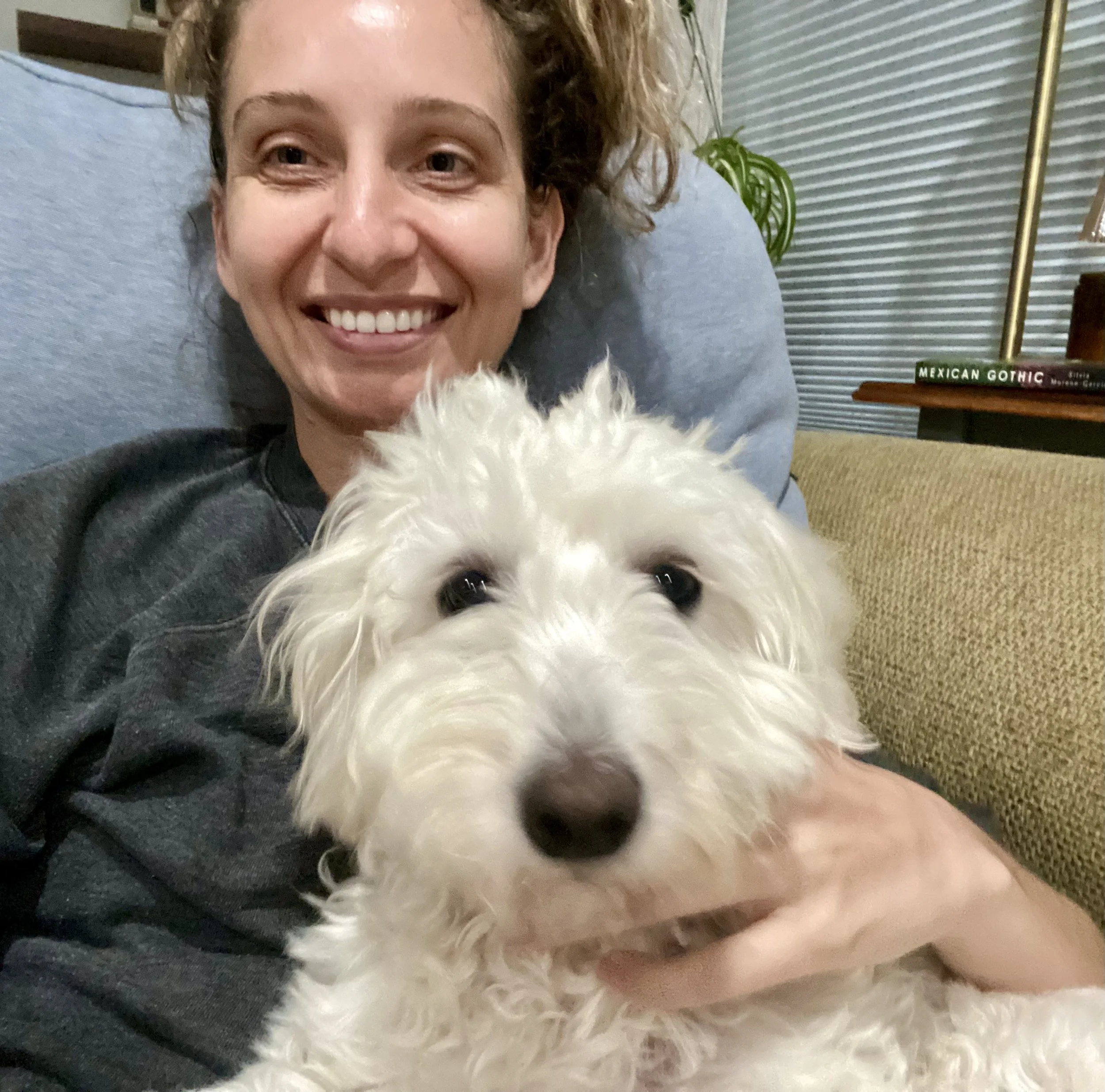 A woman with red curly hair smiling while laying on a couch, holding a fluffy white dog in front of her. In the background, there is a bookshelf with books and a tall lamp, and a table with a book titled 'Mexican Gothic' and some greenery.