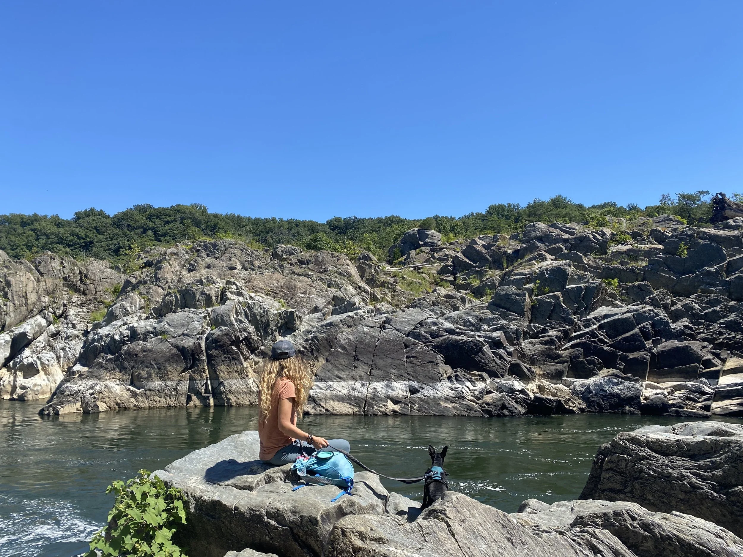 A woman with long curly blonde hair wearing a cap, sitting on rocks by a body of water, with a black dog, surrounded by trees and rocky hills under a clear blue sky.