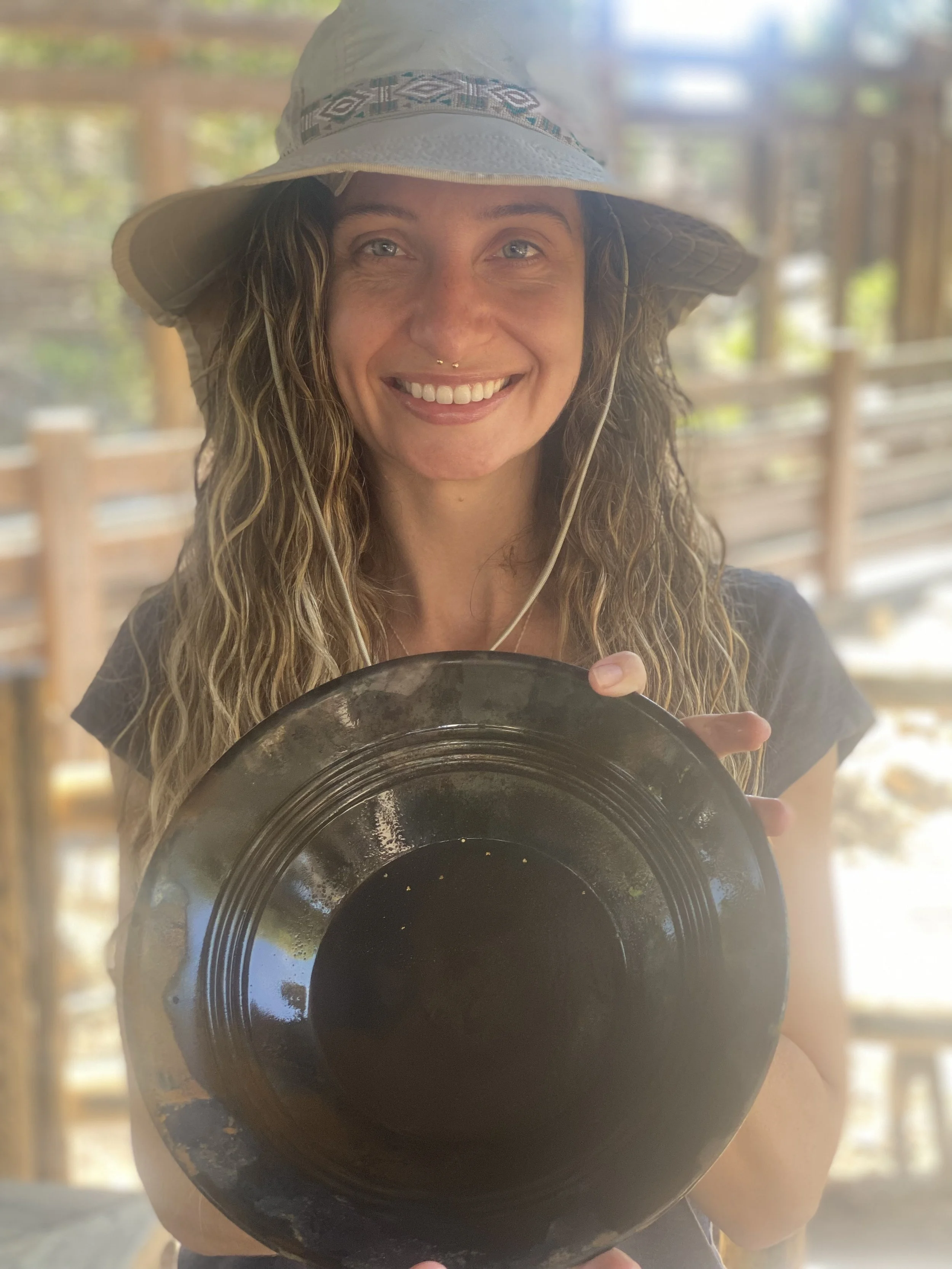 A woman with long curly hair, wearing a wide-brimmed hat, holding a dark ceremonial bowl, smiling outdoors with a wooden fence and trees in the background.